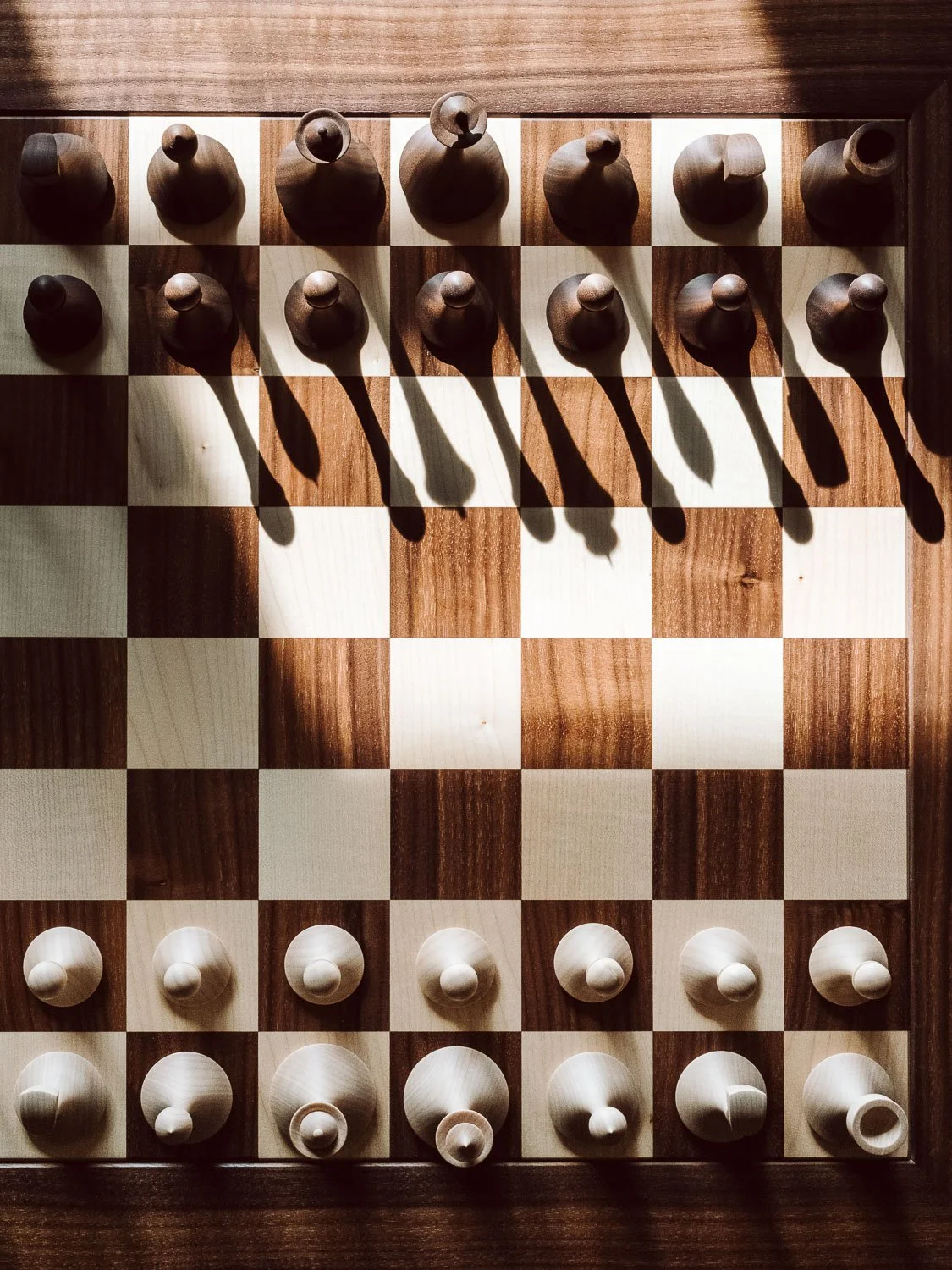 wooden chessboard set up with all the pieces in starting positions, with shadows cast by the pieces.