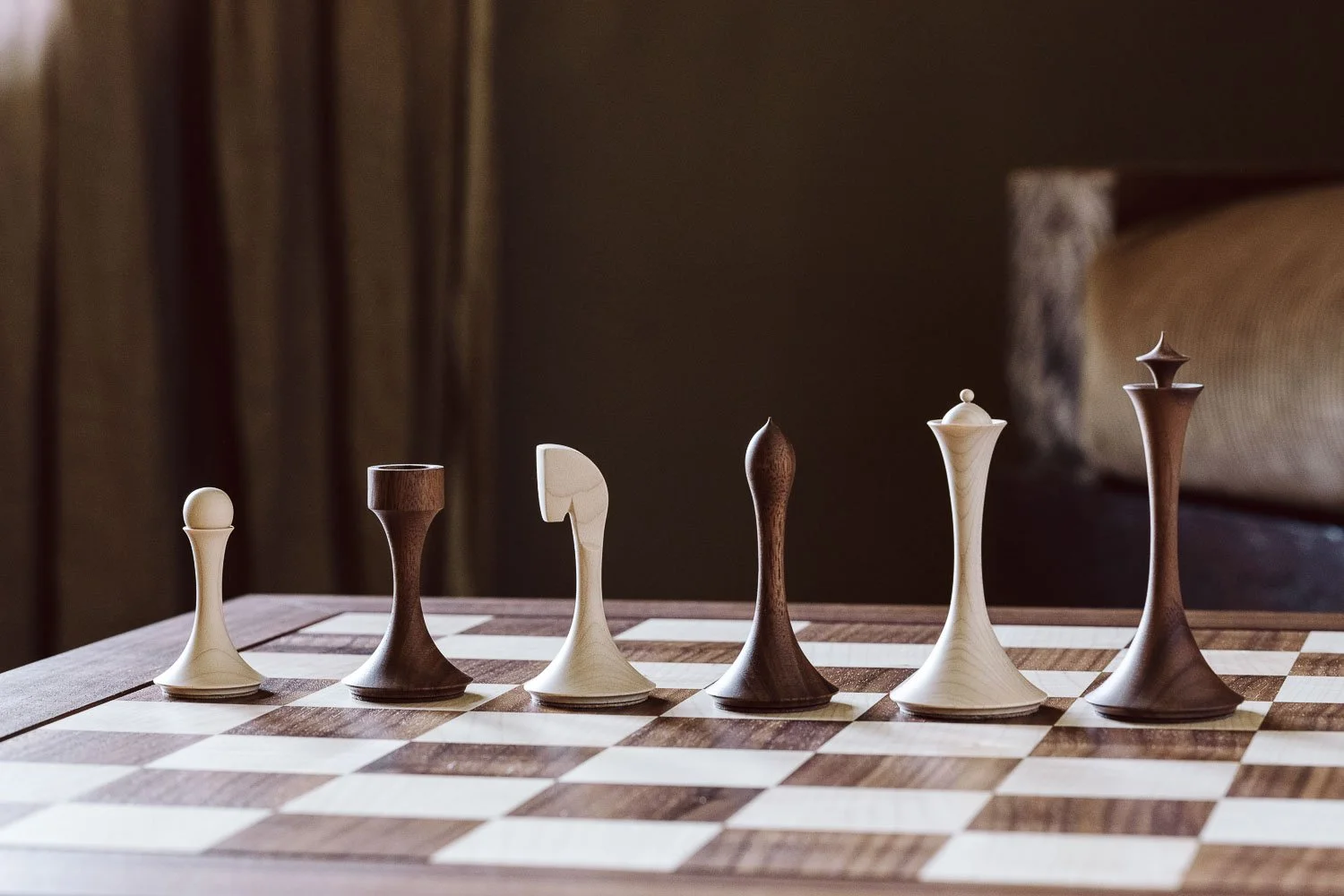 A chessboard with a row of chess pieces, including pawns and knights, made of wood in various shades from light to dark, set on a wooden table in a warmly lit room.