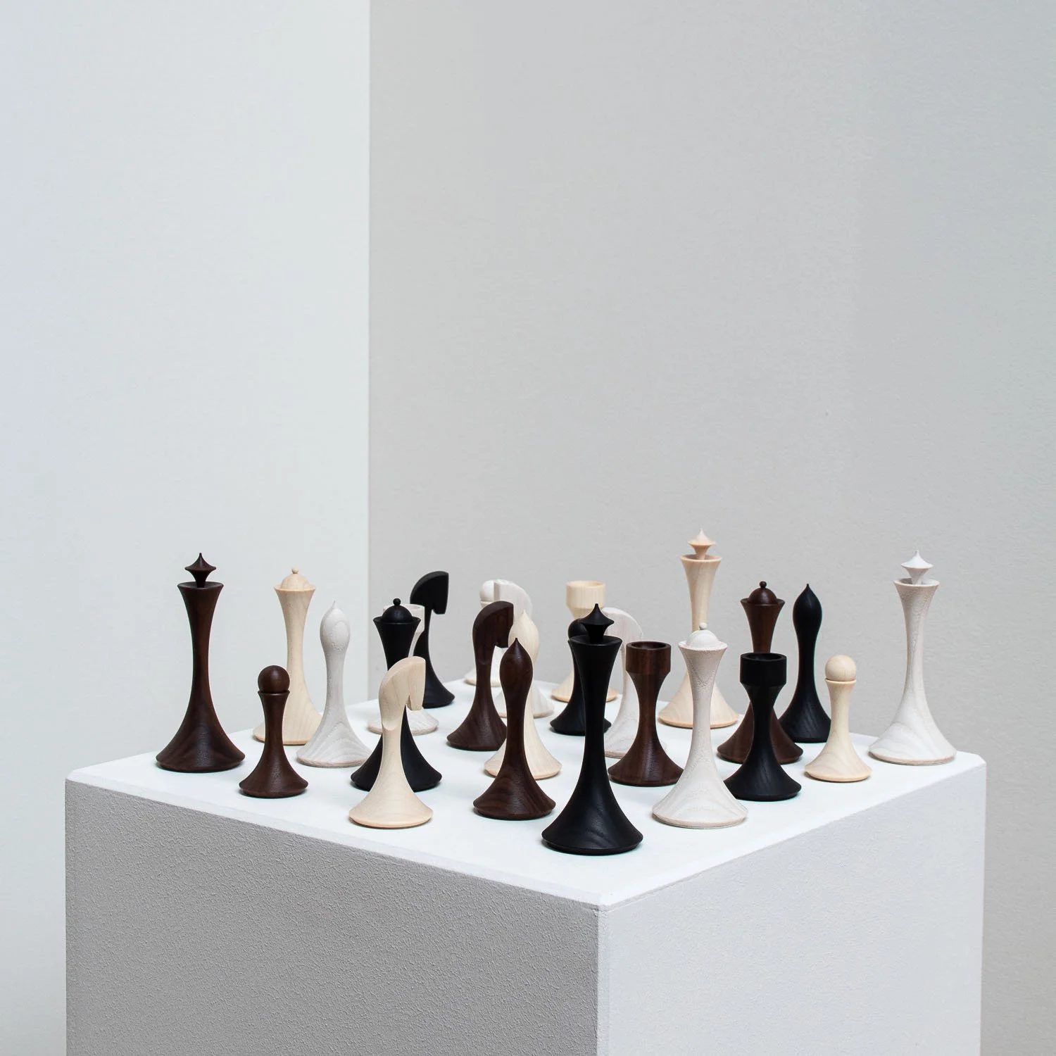 Various modern, abstract chess pieces in light, dark, and two-tone wood finishes displayed on a white pedestal against a plain background.