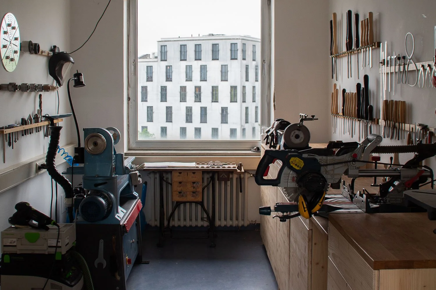 A well-organized workshop with tools on the walls, a large window letting in natural light, a workbench, and various power tools including a miter saw and a drill.