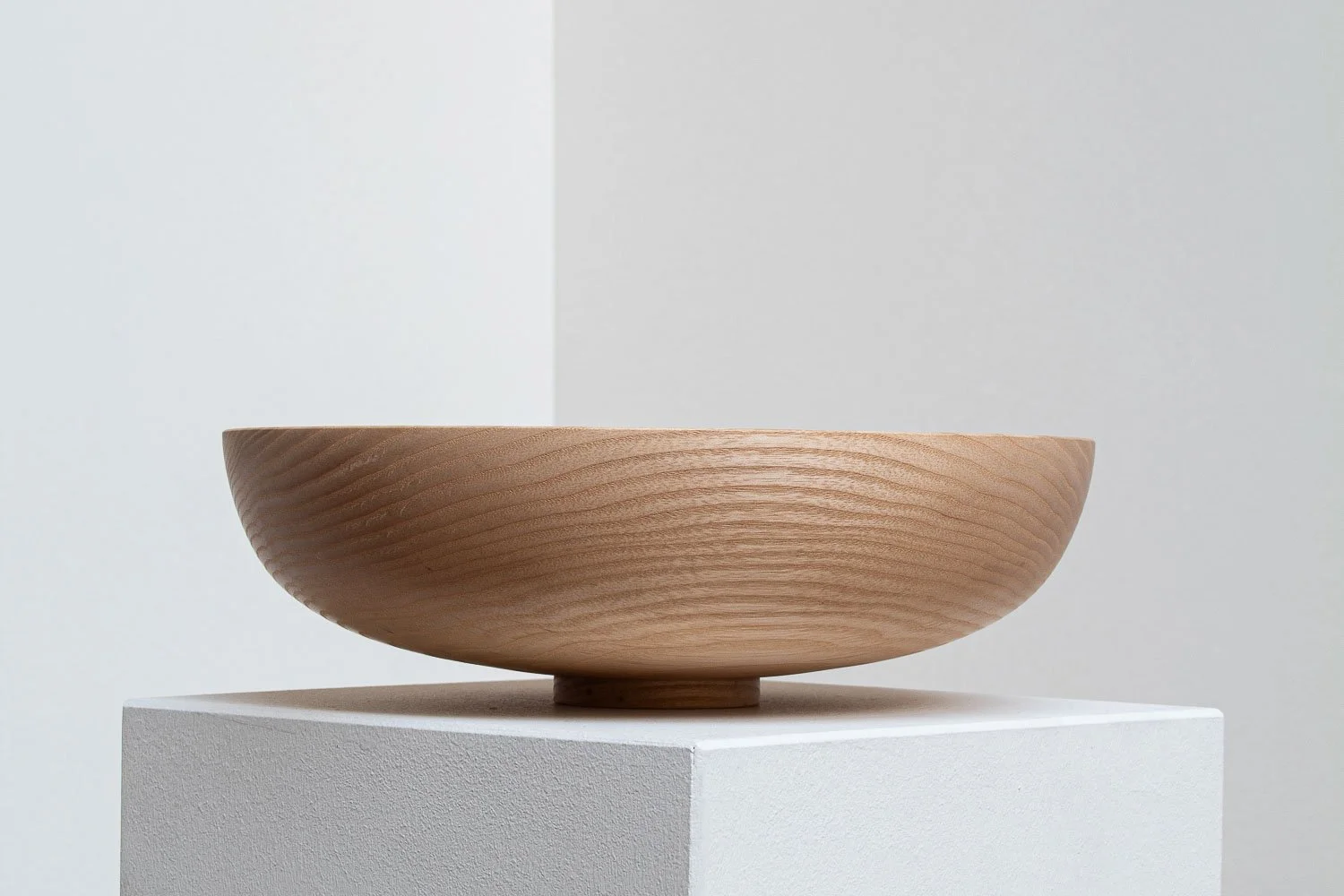 A minimalist wooden bowl resting on a white pedestal against a plain, neutral background.