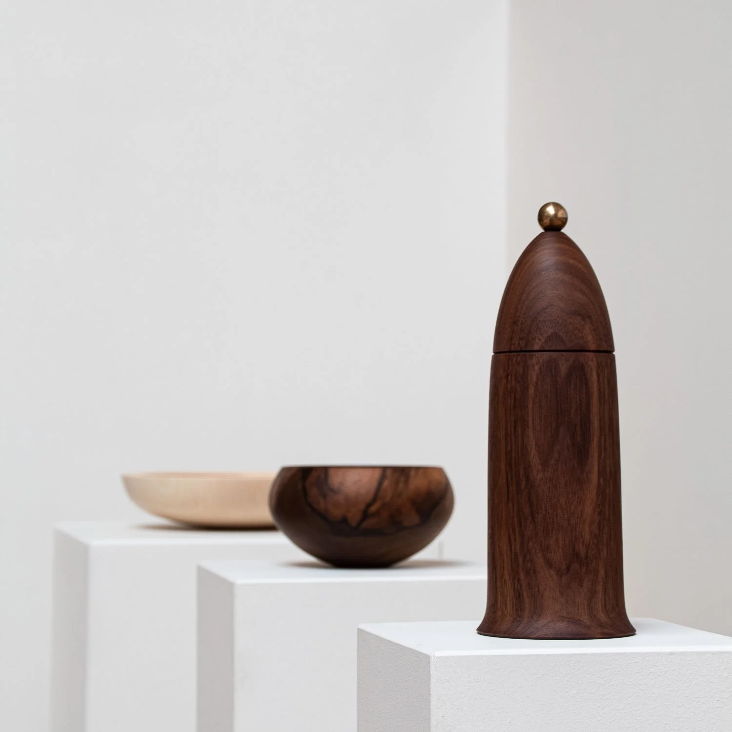 Wooden and ceramic bowls and a wooden sculpture displayed on white pedestals in an art gallery.