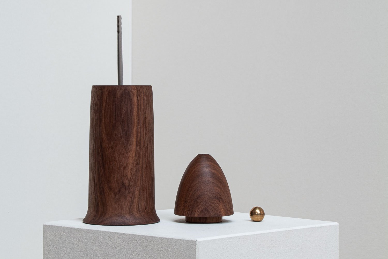 Three modern decorative objects made of wood and metal on a white pedestal, including a tall wooden cone, a smaller wooden dome, and a small metallic sphere, against a light gray background.