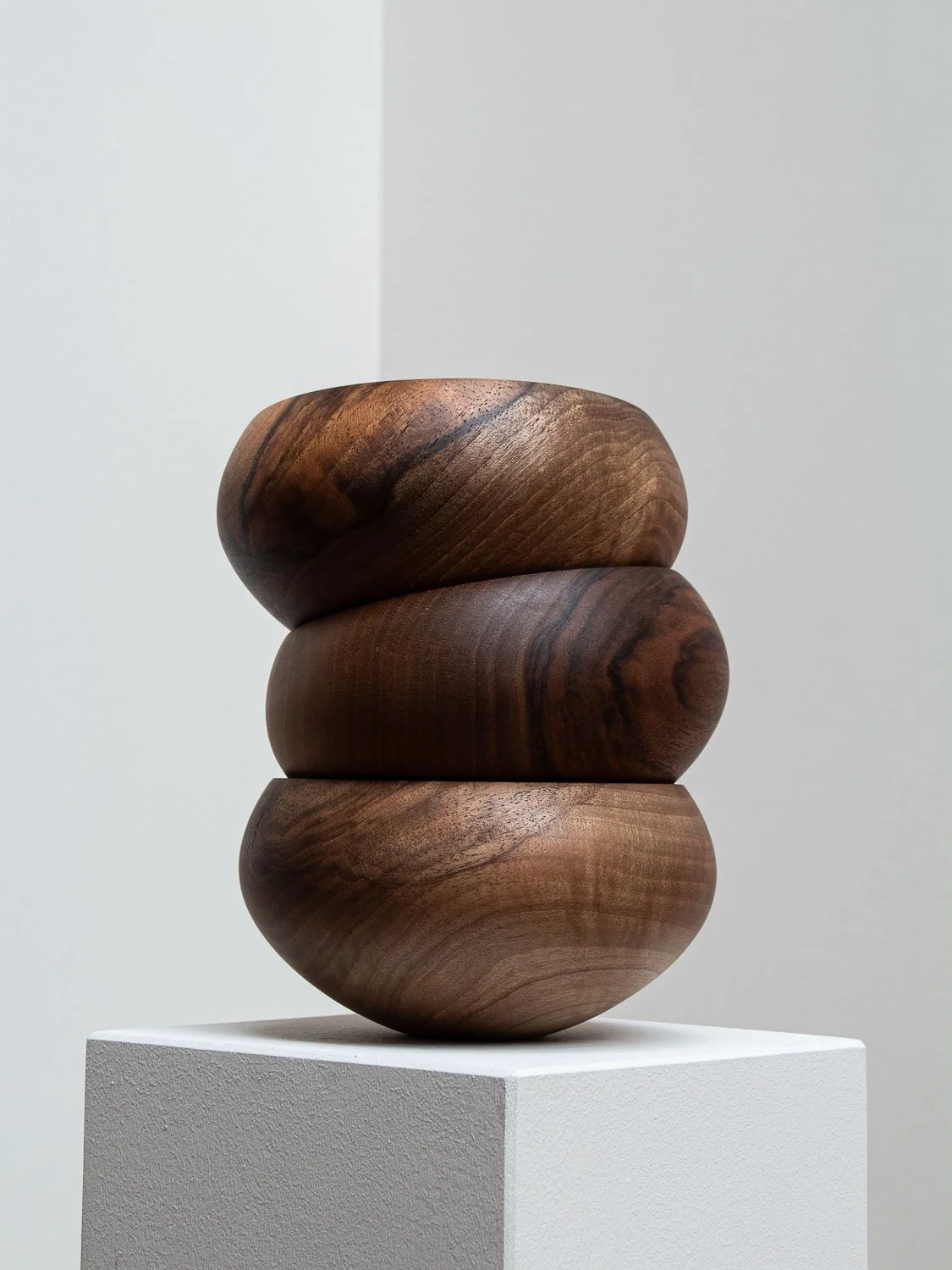 A sculpture made of three polished, oval-shaped wooden pieces stacked on a white pedestal against a gray and white background.