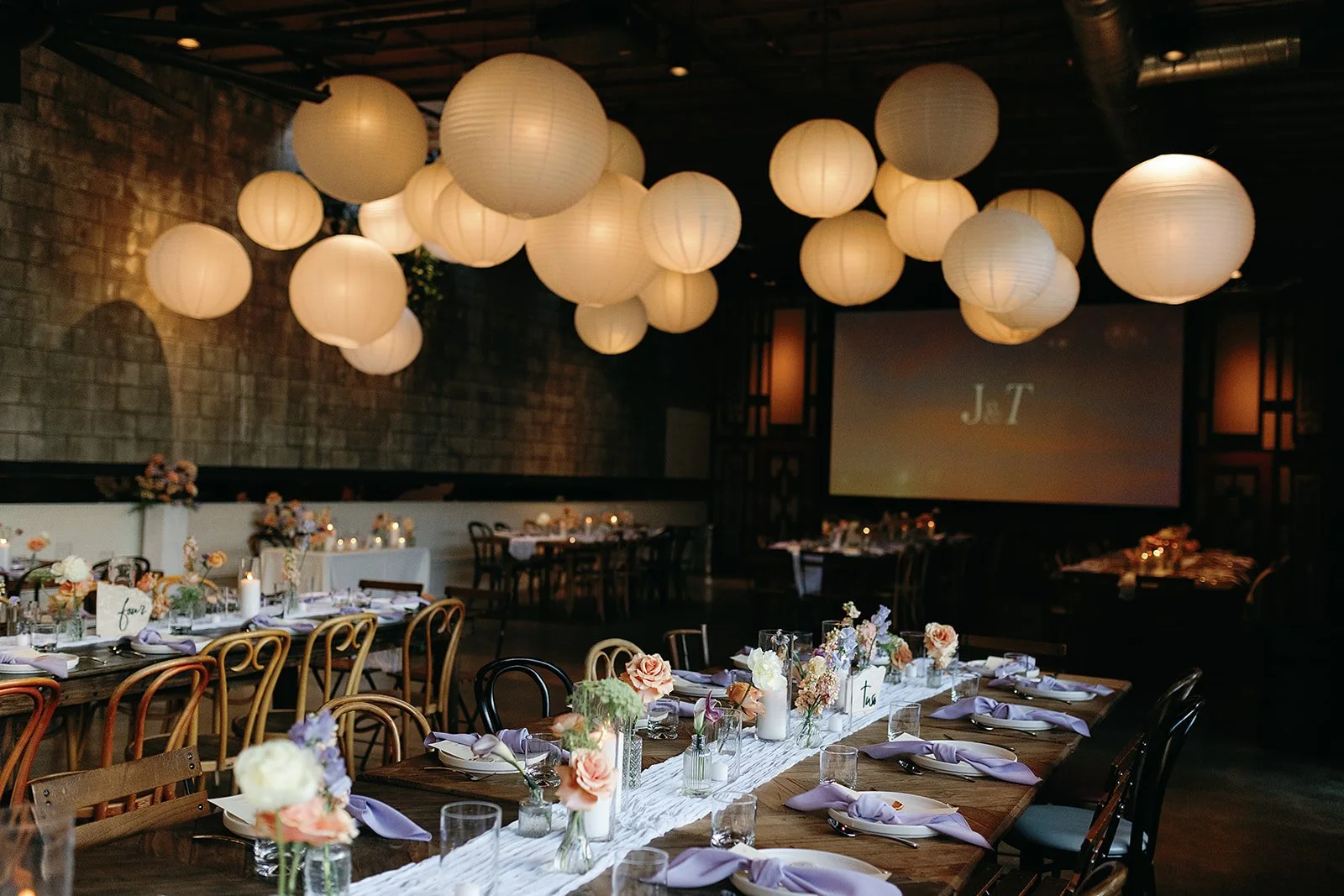 Wedding reception setup with long tables decorated with floral centerpieces, candles, and lavender napkins, hanging white paper lanterns overhead, and a large screen displaying "J + T" in the background.