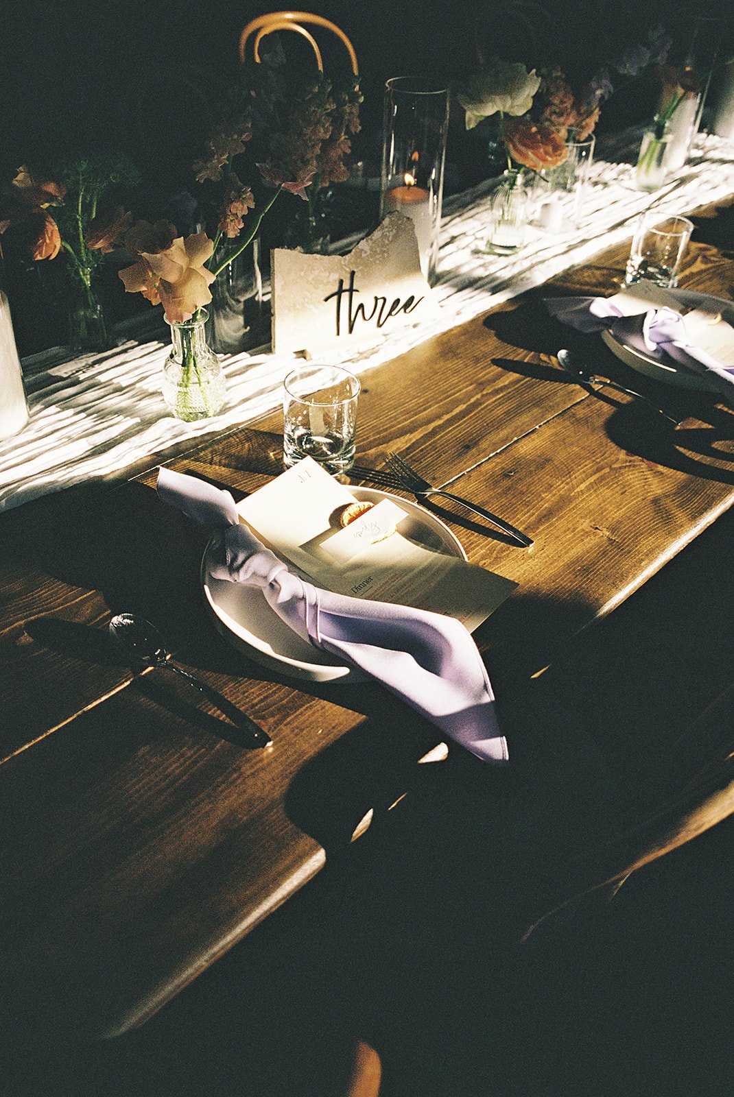 A close-up of a wedding reception table setting with a wooden table, white plates with lavender napkins, clear glassware, and a menu. Flowers in vases and a table sign with the word 'three' are visible in the background. The setting appears elegant a