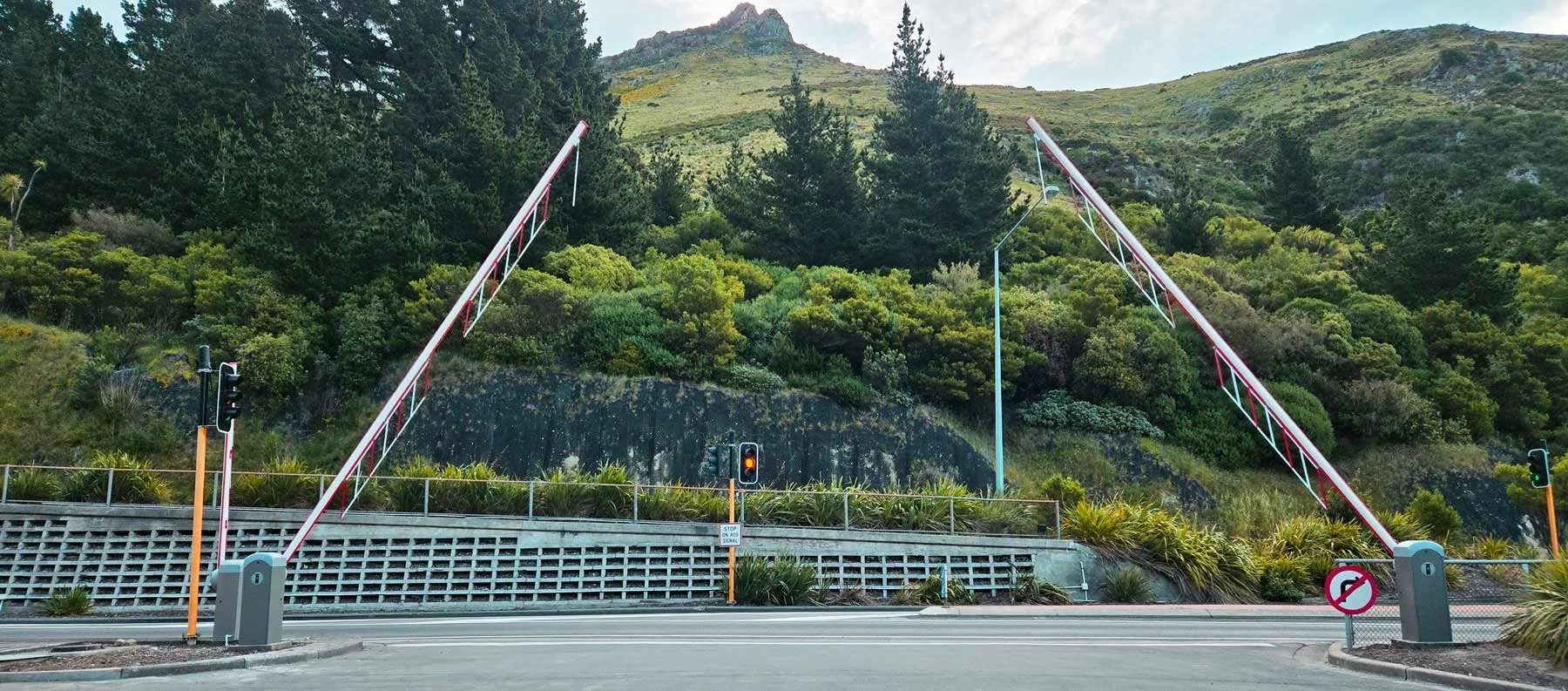 Red and white striped barrier arms across a road, traffic lights, and greenery with trees and a mountain in the background.