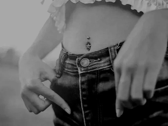 Close-up of a person wearing high-waisted jeans and a ruffled crop top, showing their midriff with a belly button piercing and a decorative gemstone.