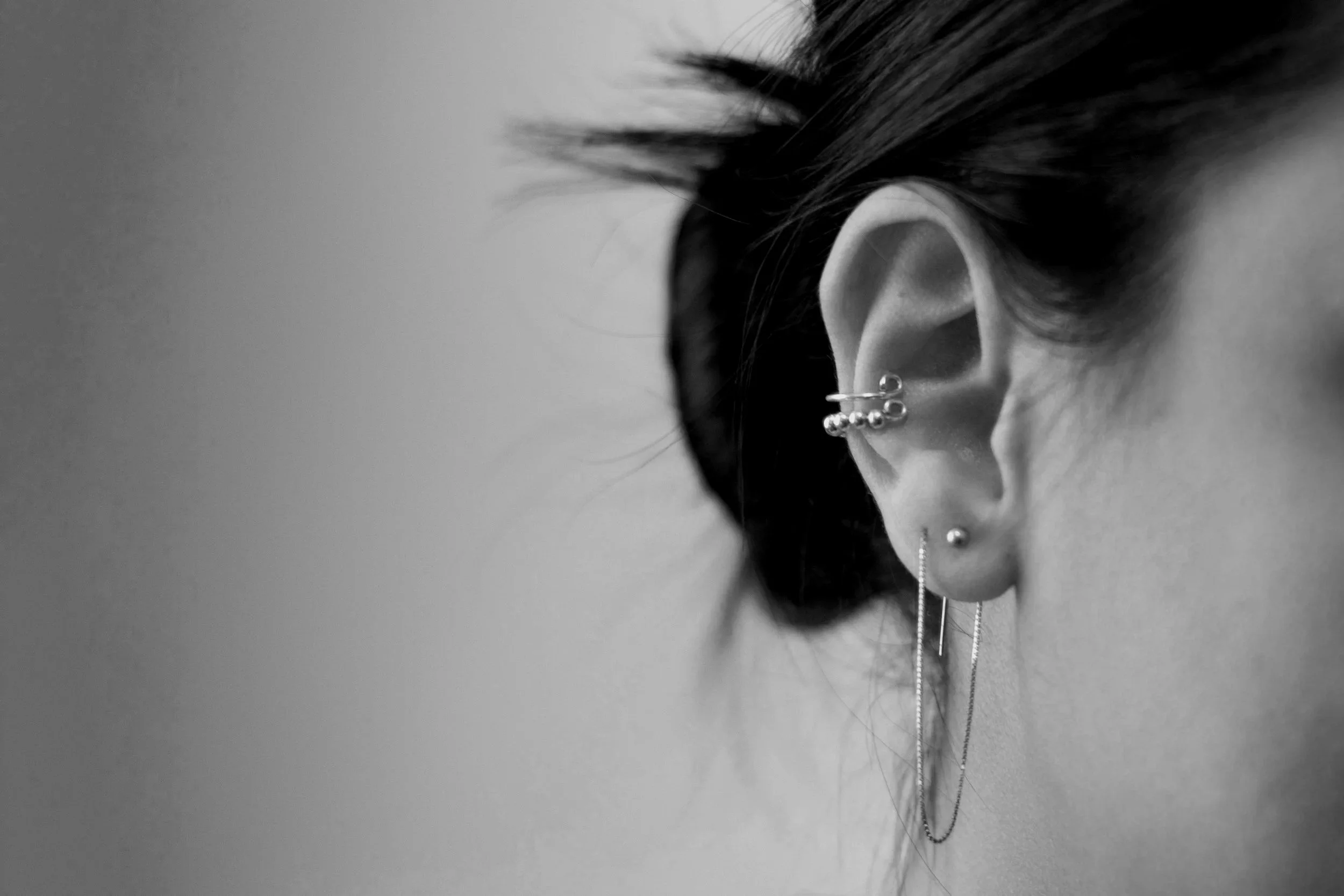 Close-up black and white photo of a person's ear with multiple silver earrings, including a helix cuff, a cartilage ball stud, and a dangling chain earring.
