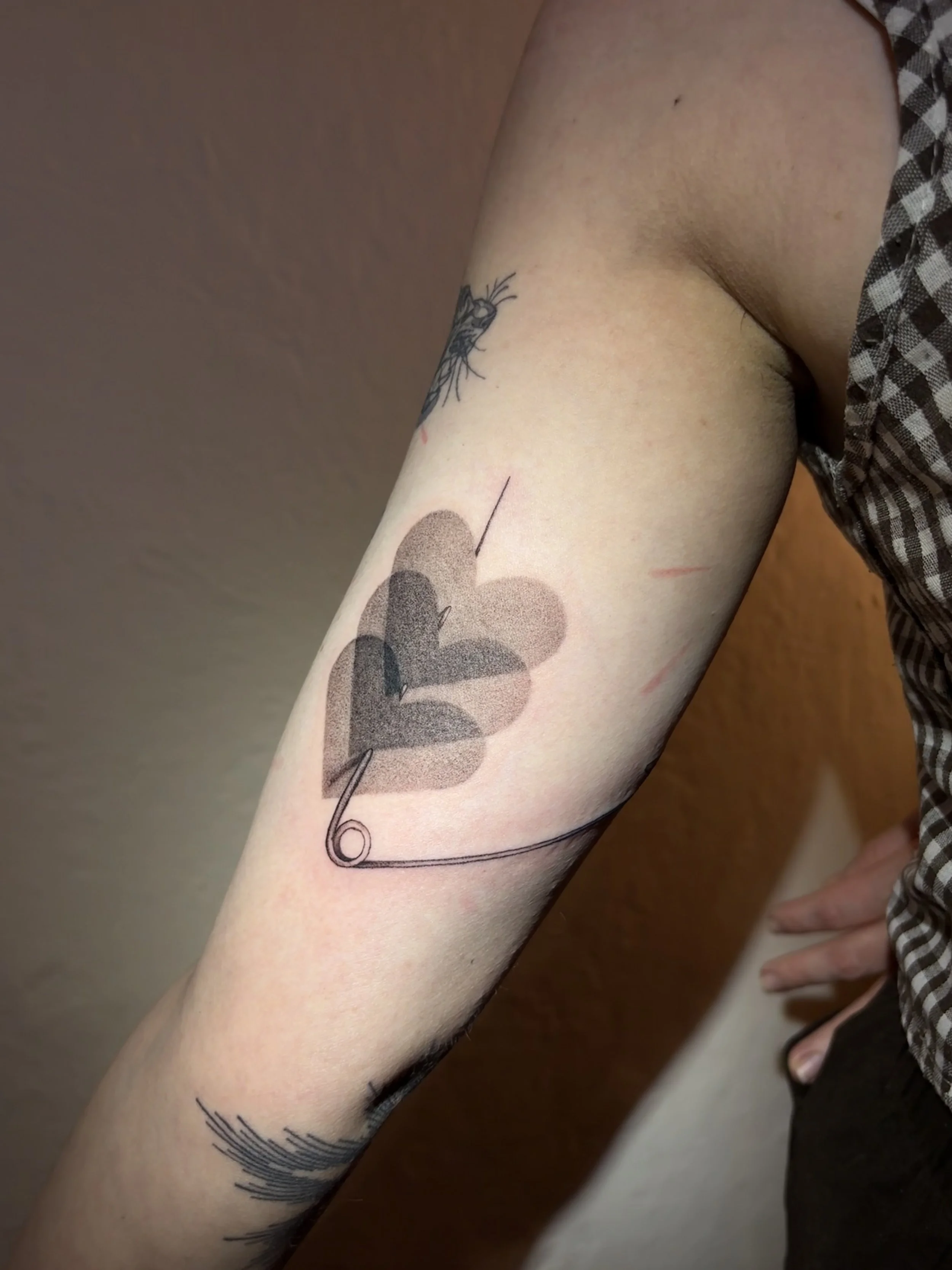 Close-up of a person's arm with a tattoo of a heart, some abstract shapes, and a safety pin design.