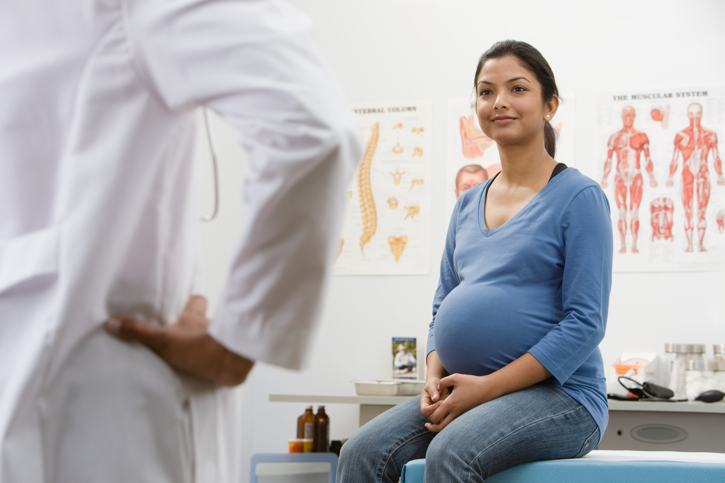 A pregnant woman with dark hair sits on an examination table while speaking with a doctor in a white coat.