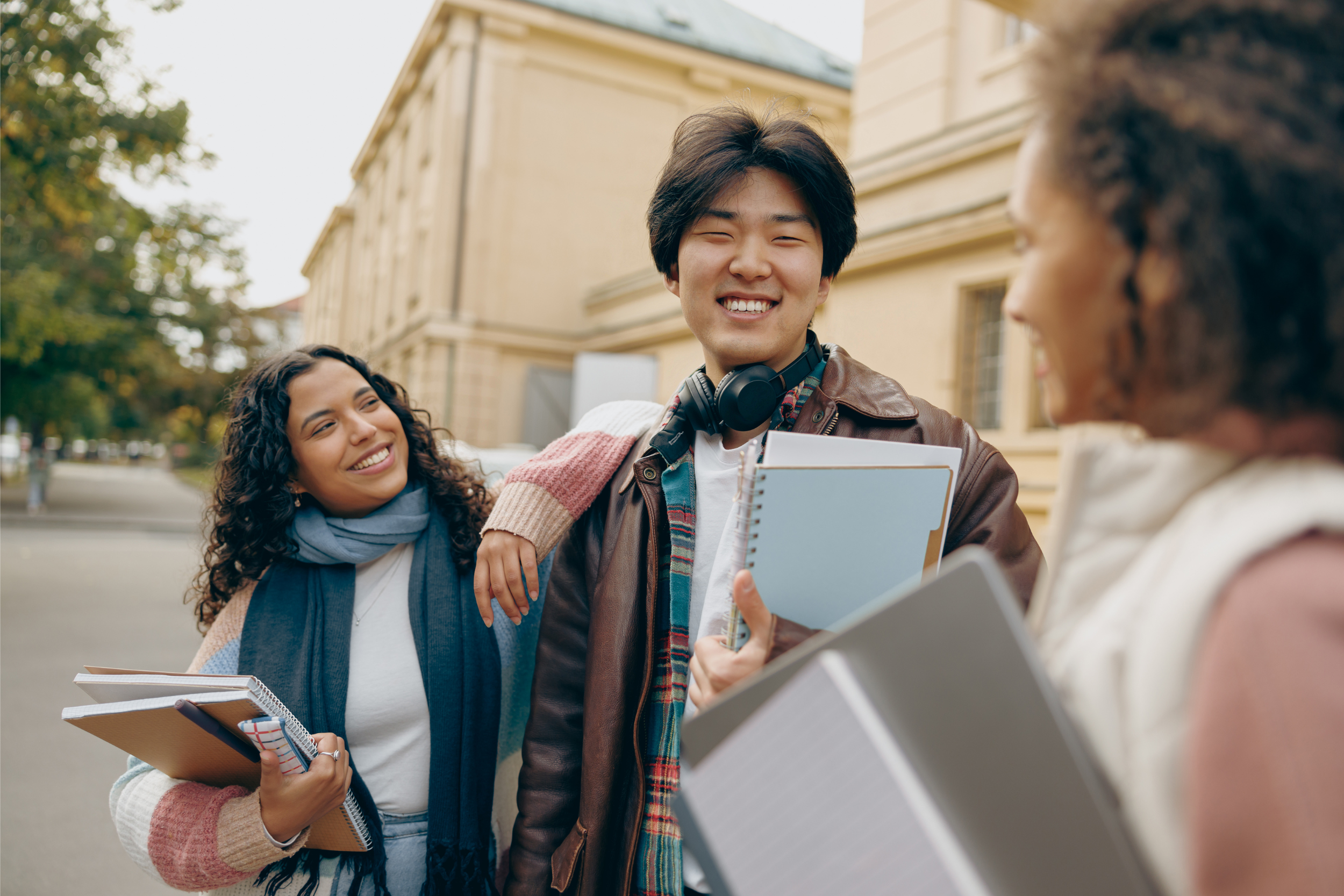 A group of three international students standing outside of a university, chatting and smiling.