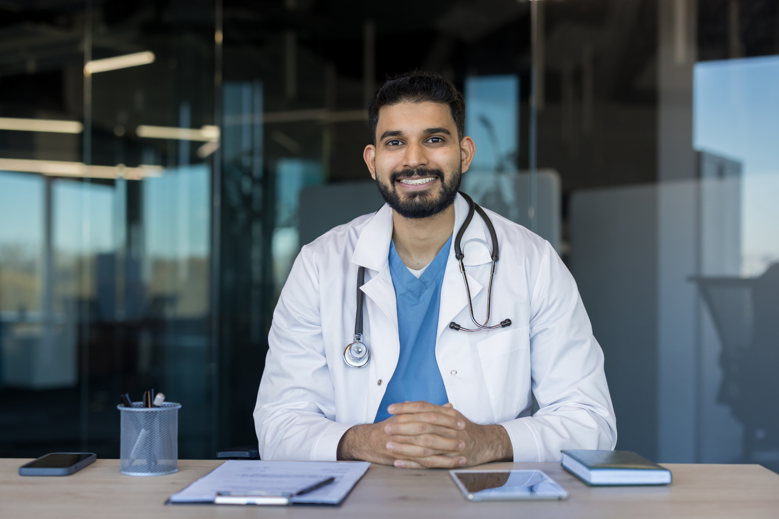 A smiling male doctor, sitting at a desk with a clipboard and tablet on the table in front of him.