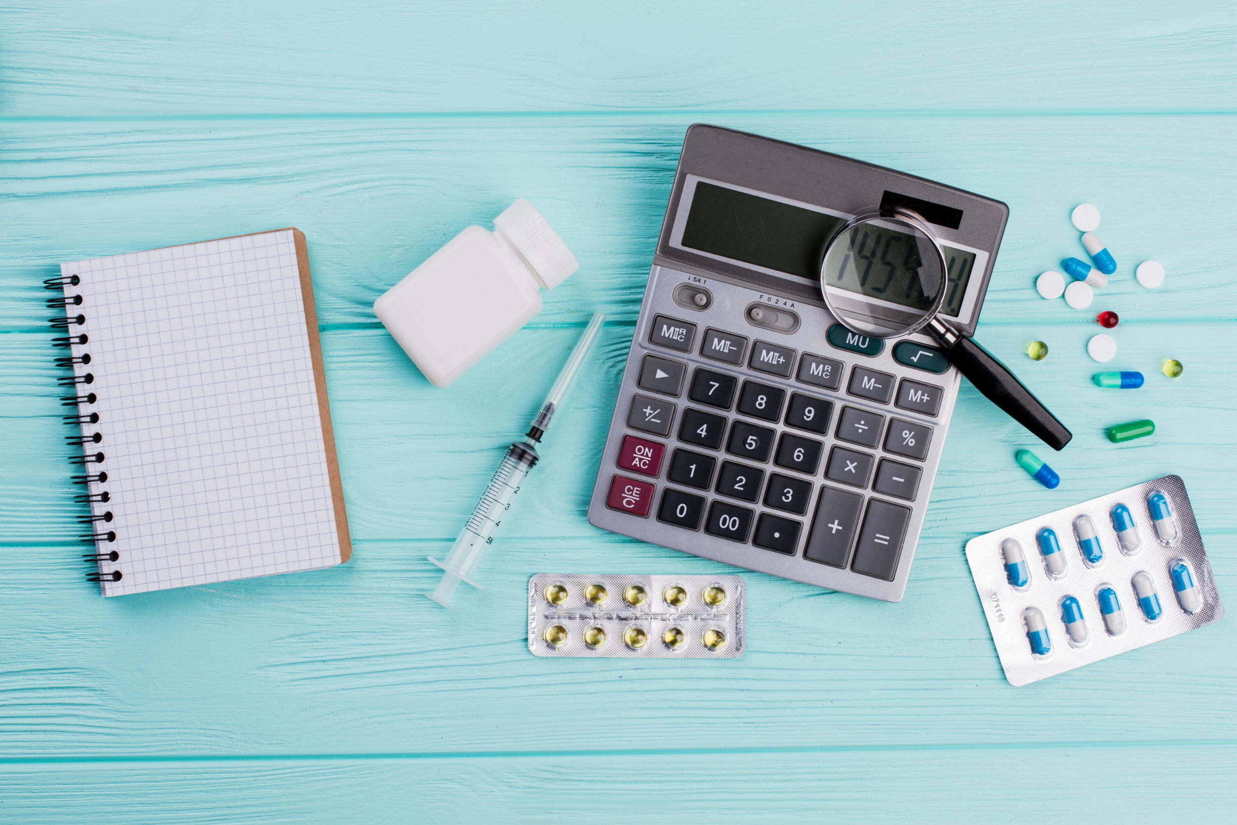 Graph book, pill bottle, pills, a needle, a calculator, and a magnifying glass on a light blue wood background.