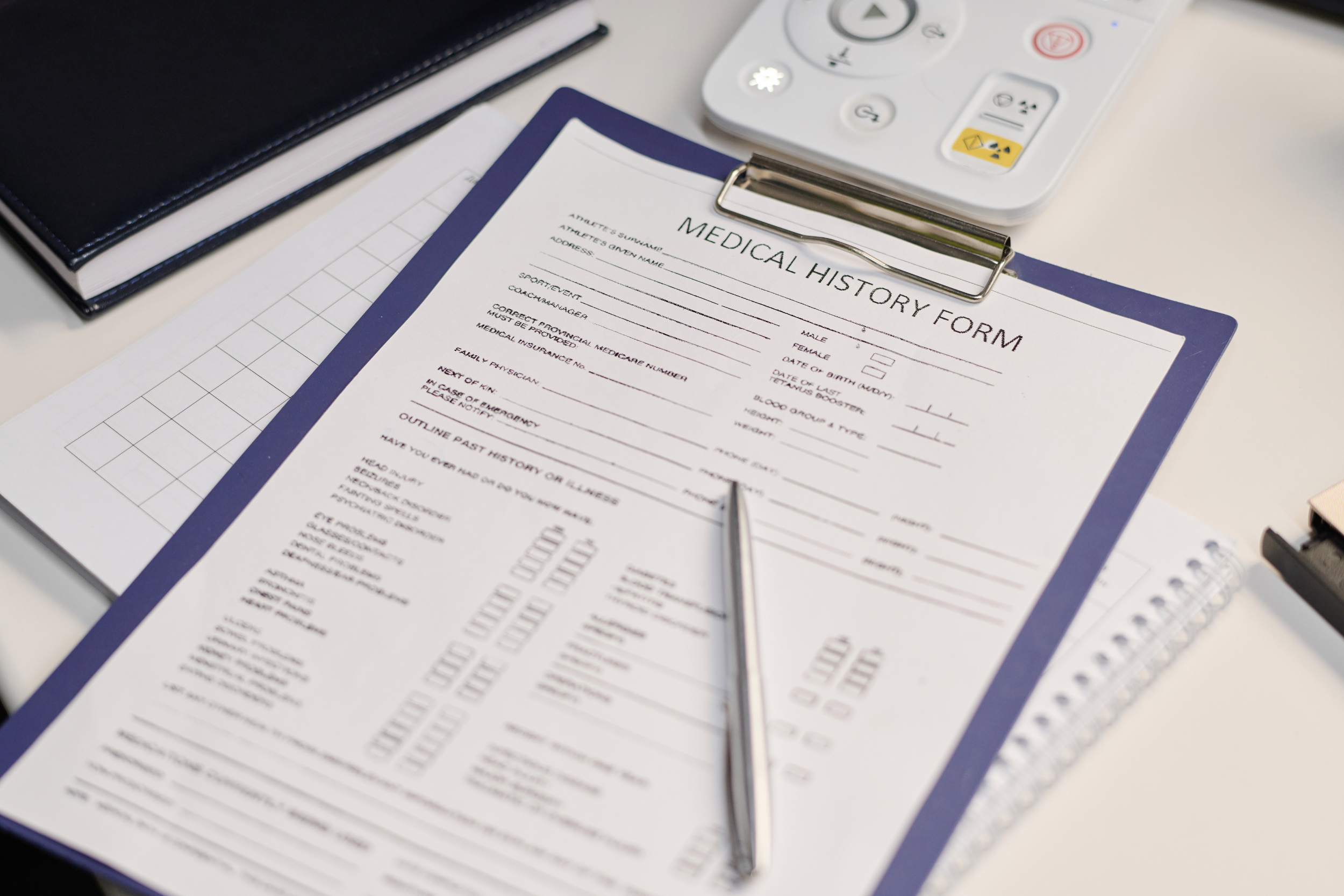 A medical history form on a blue clipboard sits on a desk. A silver pen is on top of the form.