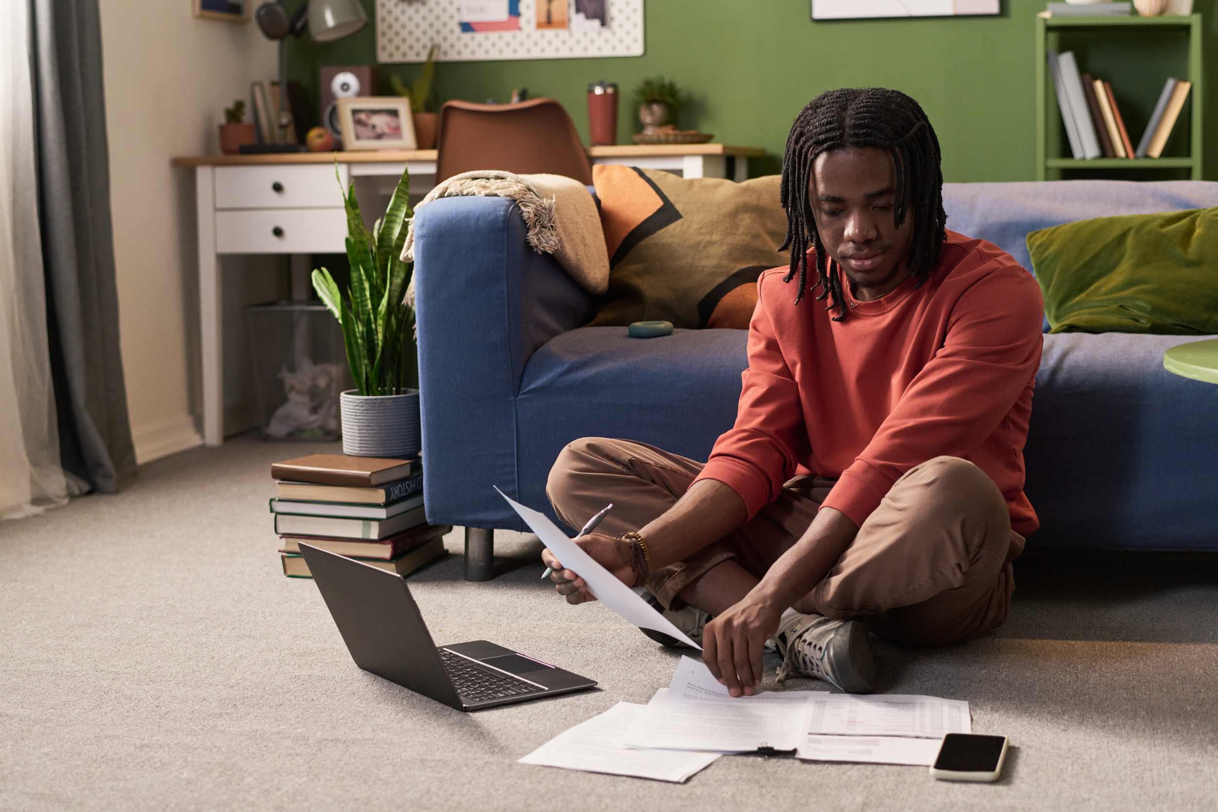 A young man sitting on the floor, reviewing documents with an open laptop in front of him.