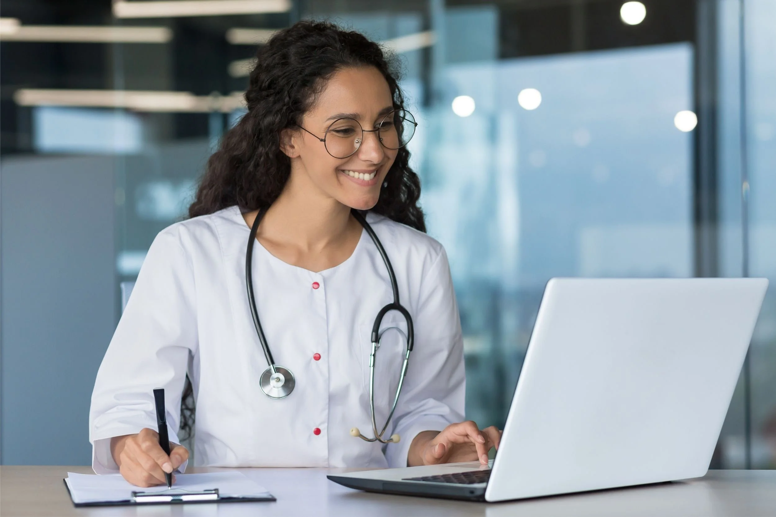 A doctor types on a laptop while double-checking info from a clipboard.