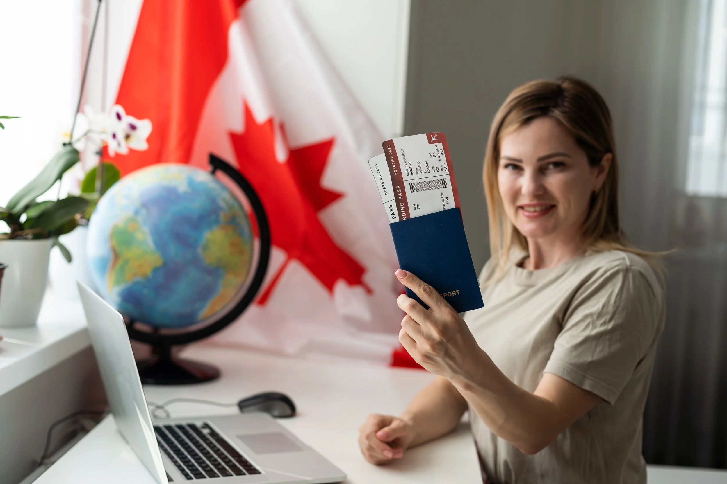 Woman with passport and plane tickets sitting at laptop with Canadian flag in background to represent checking CRS score and readiness to apply for permanent residency