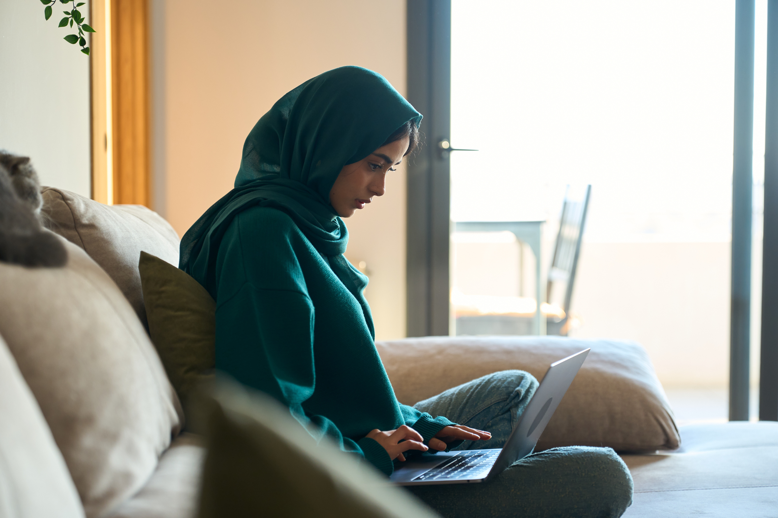 Young hijabi woman sitting on a couch, looking down at a laptop.