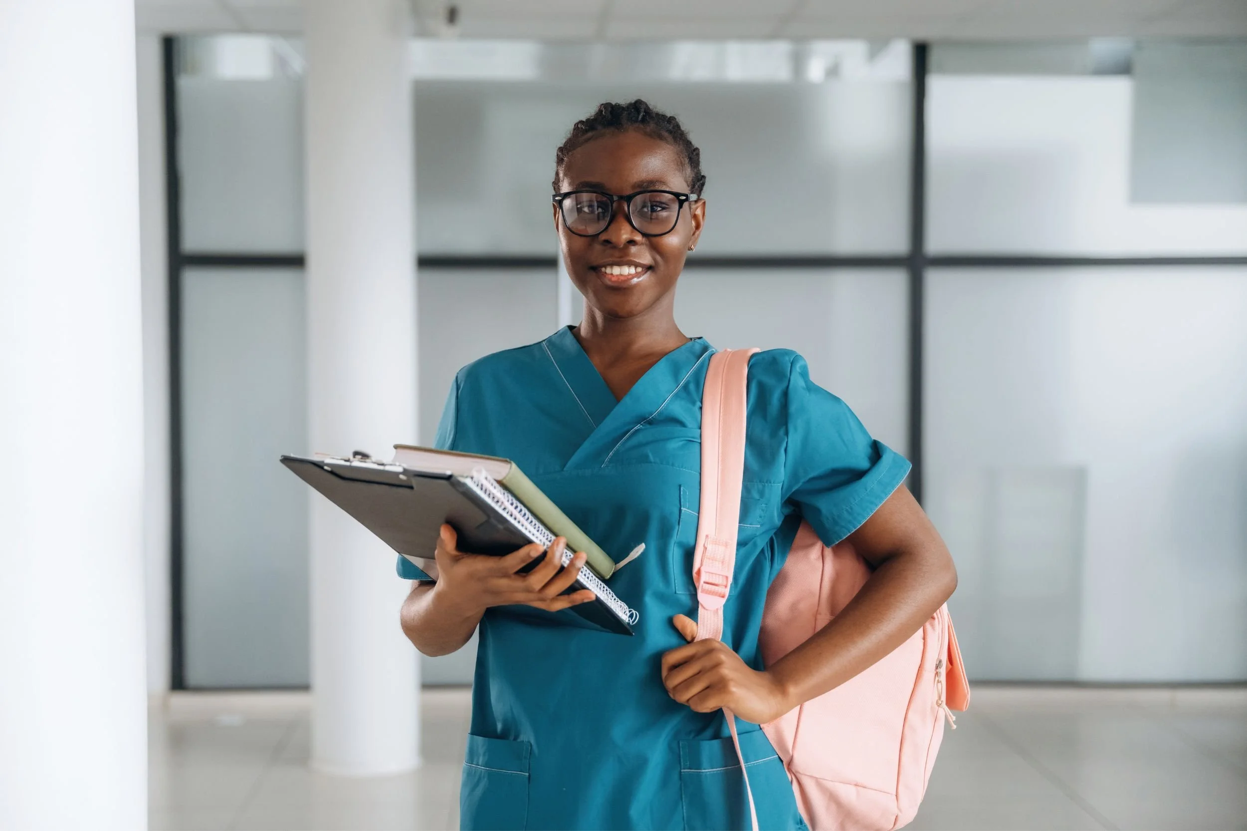 A nursing student in blue scrubs holding a pink backpack and books in a hospital.