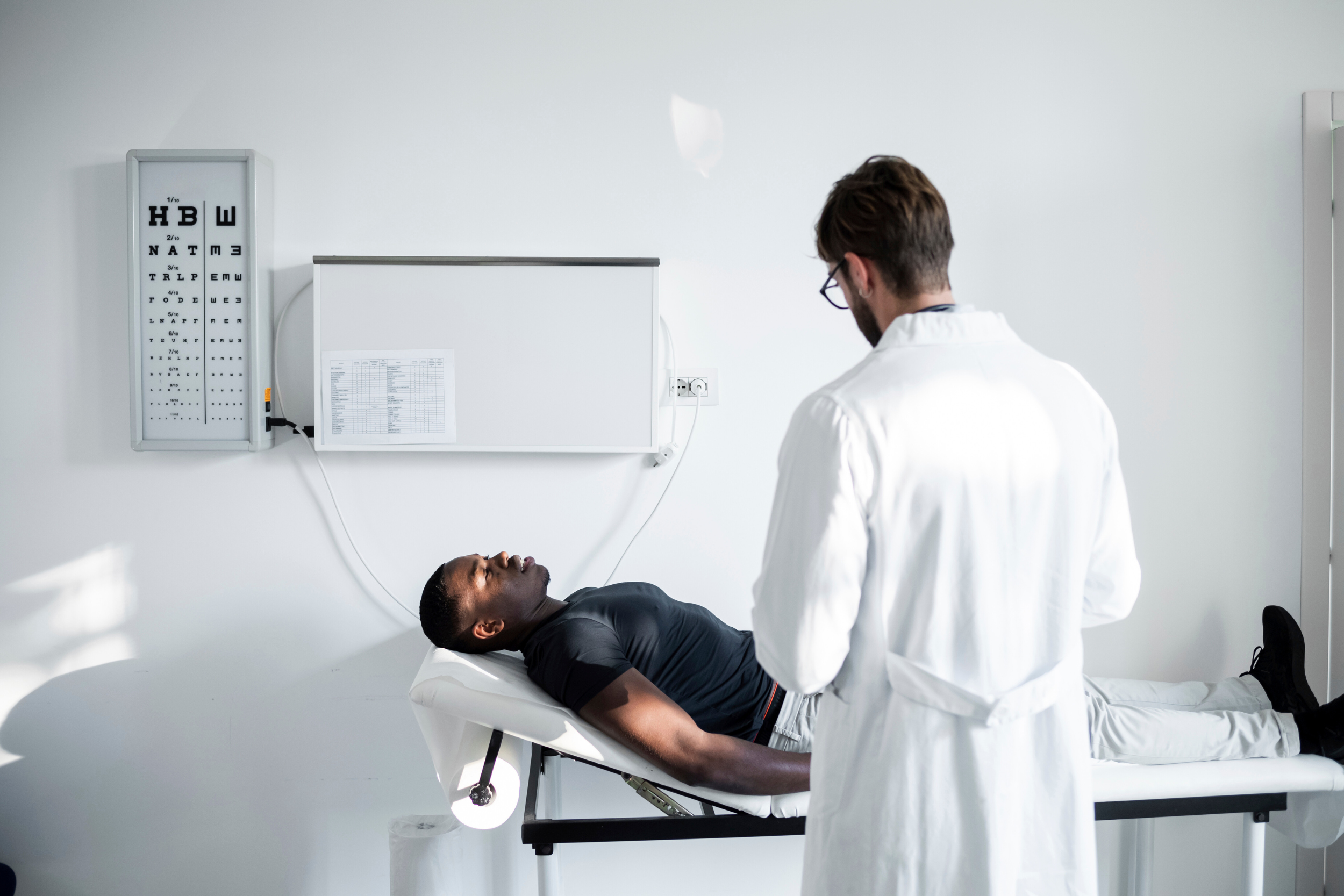 A man laying on a medical bed in an exam room, speaking with a doctor.