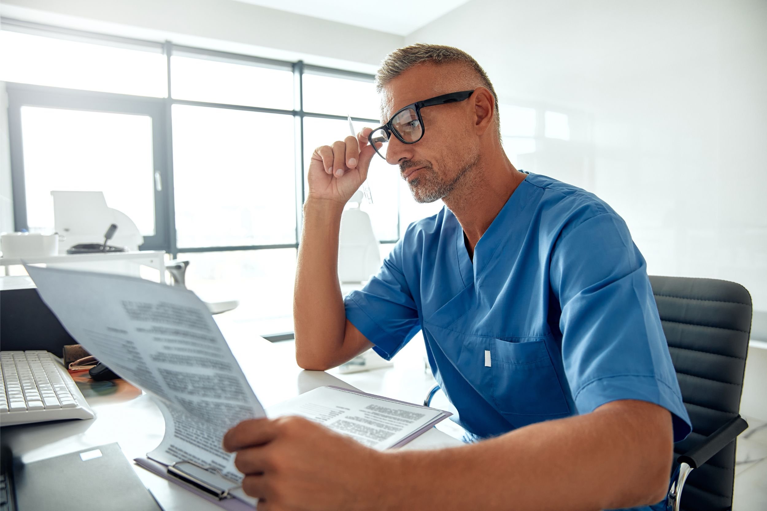 A doctor in a blue scrub shirt and black-rimmed glasses sits at a desk reviewing medical forms.