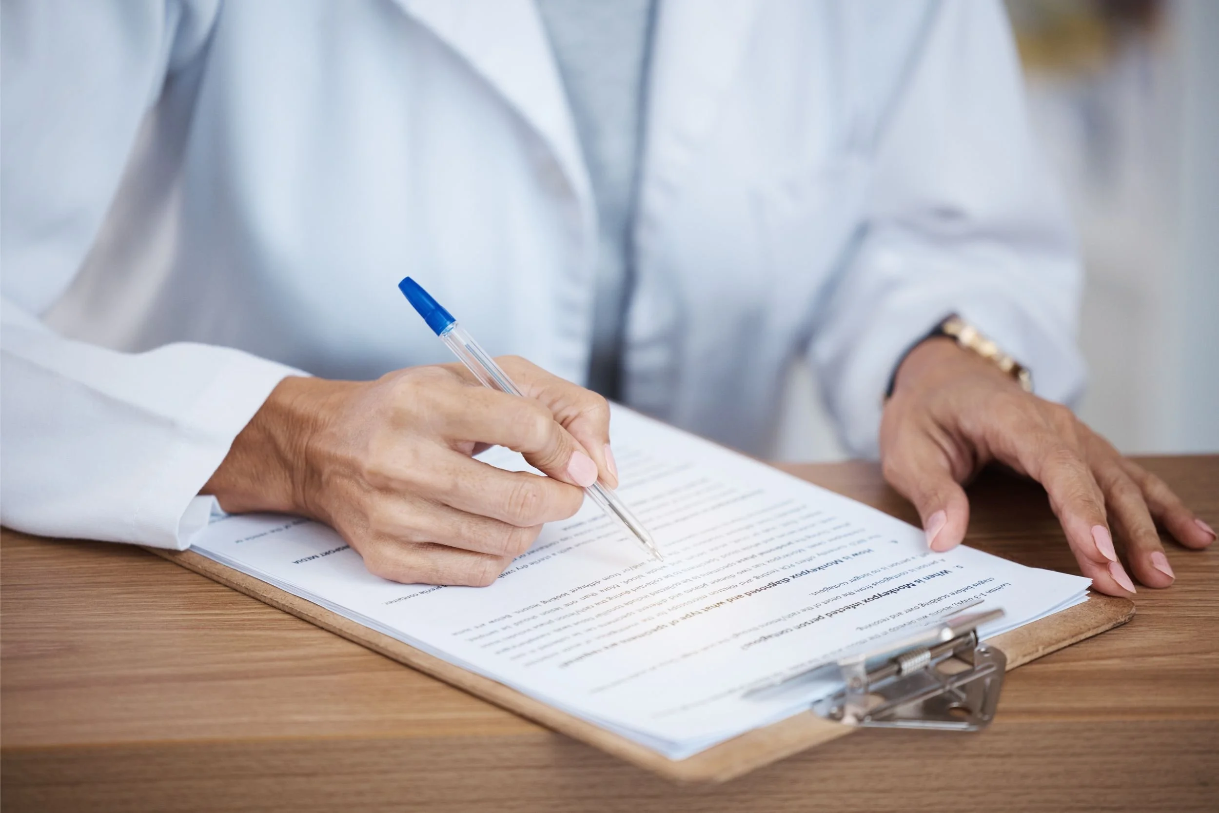 A doctor fills out medical forms on a clipboard on a desk in a clinic.