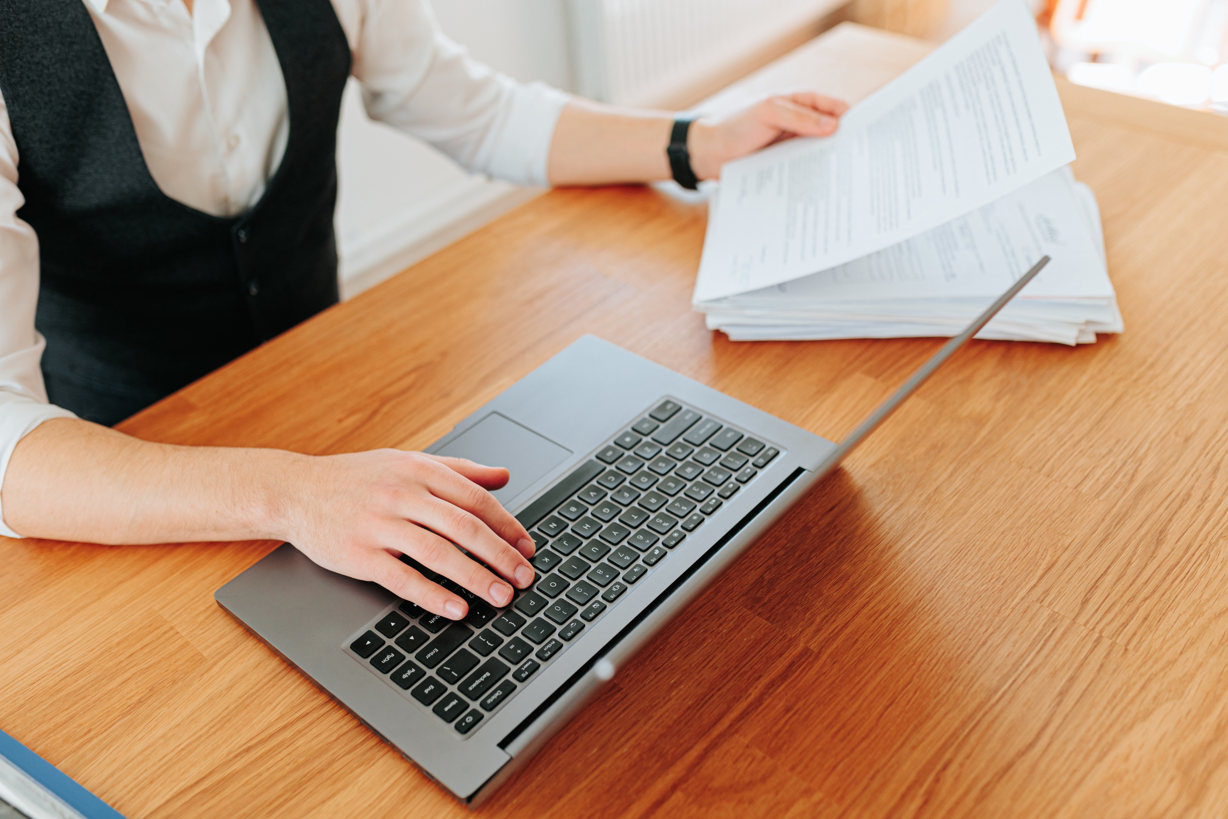 A man sitting at a desk with one hand on a laptop keyboard, reviewing paper documents in his other hand