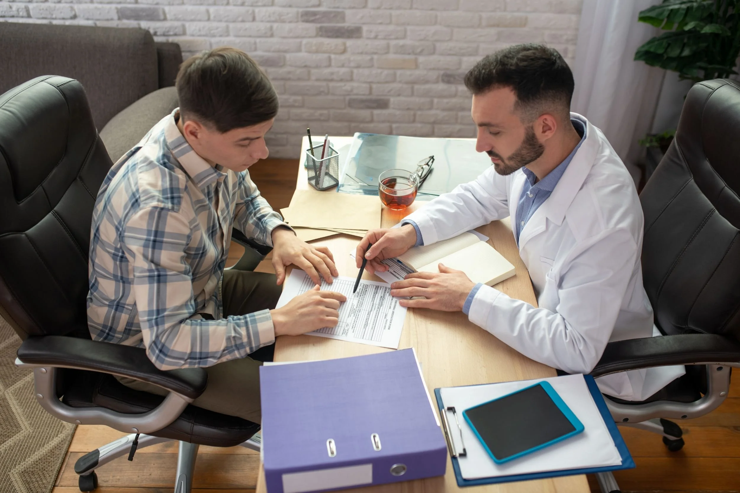 A man in a plaid shirt discusses medical documents with his physician in the physician's office. The doctor points to something on a paper with his pen.