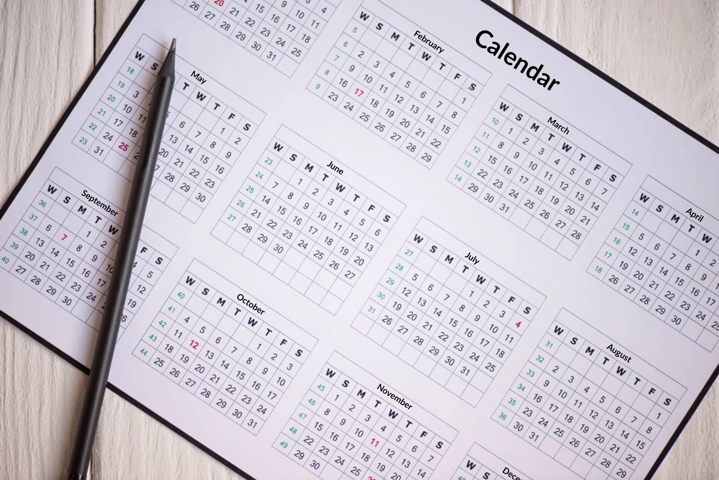A 12-month calendar lays flat on a light wooden table with a pen on it, showing the months from January through December.