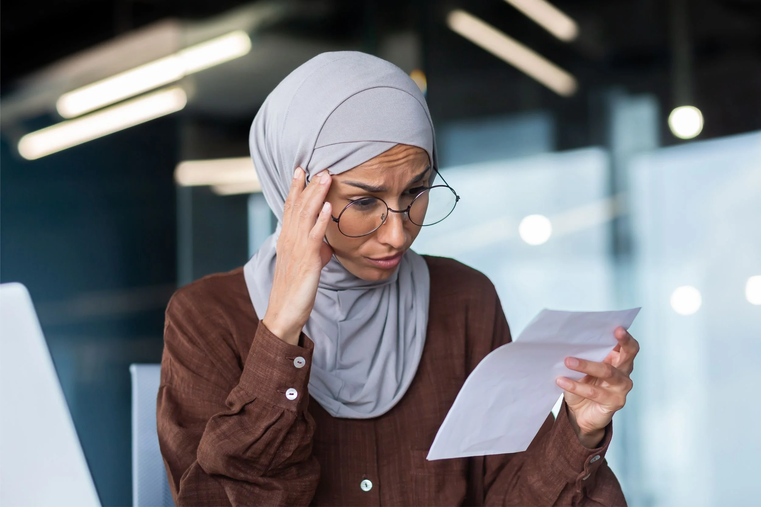 A woman wearing glasses, a brown blouse, and a grey hijab looks concerned reading a document in her hand/