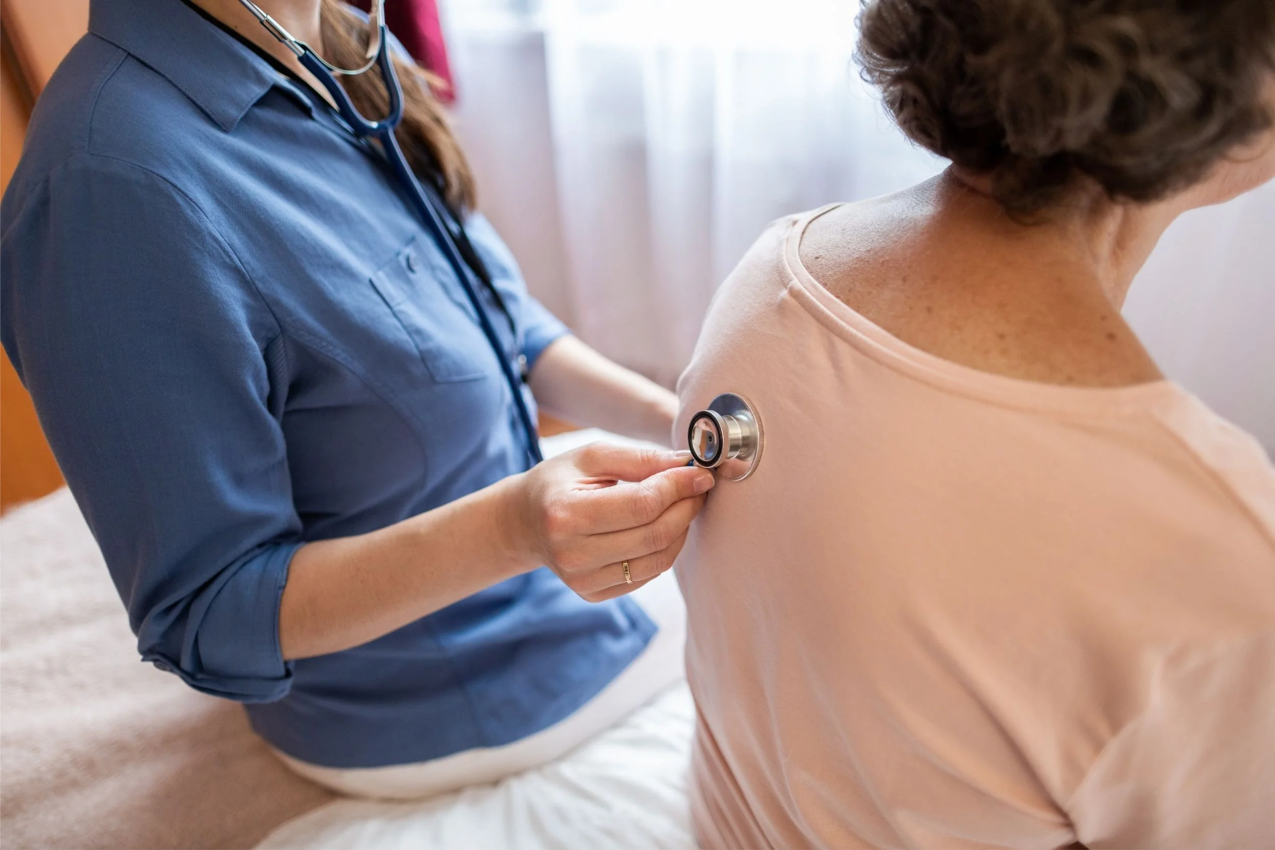 A physician holds a stethoscope to the back of an elderly patient, listening to their lungs.