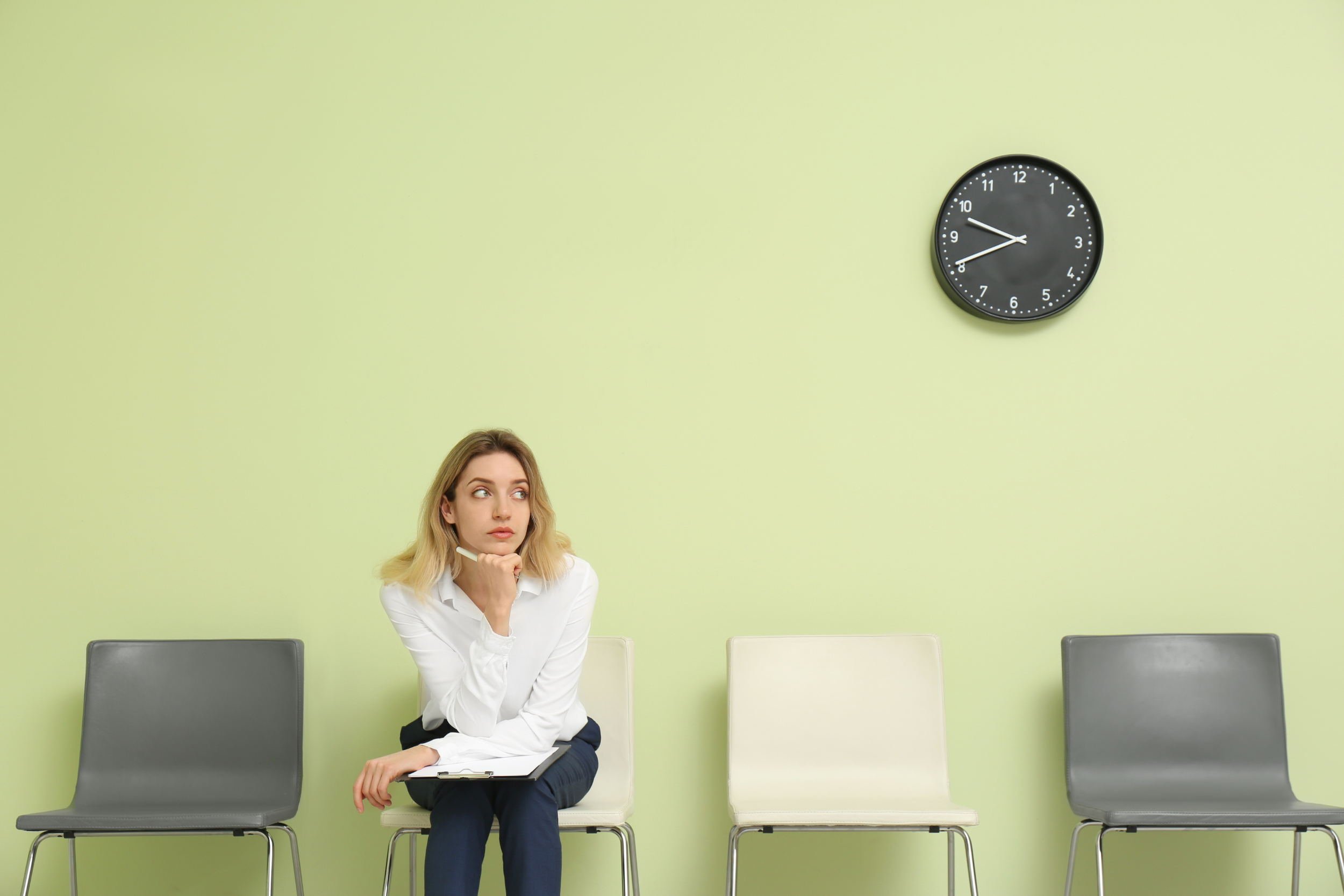 A young woman sits in a waiting room, clipboard in her lap, chin in her hand. There's a large clock on the wall.