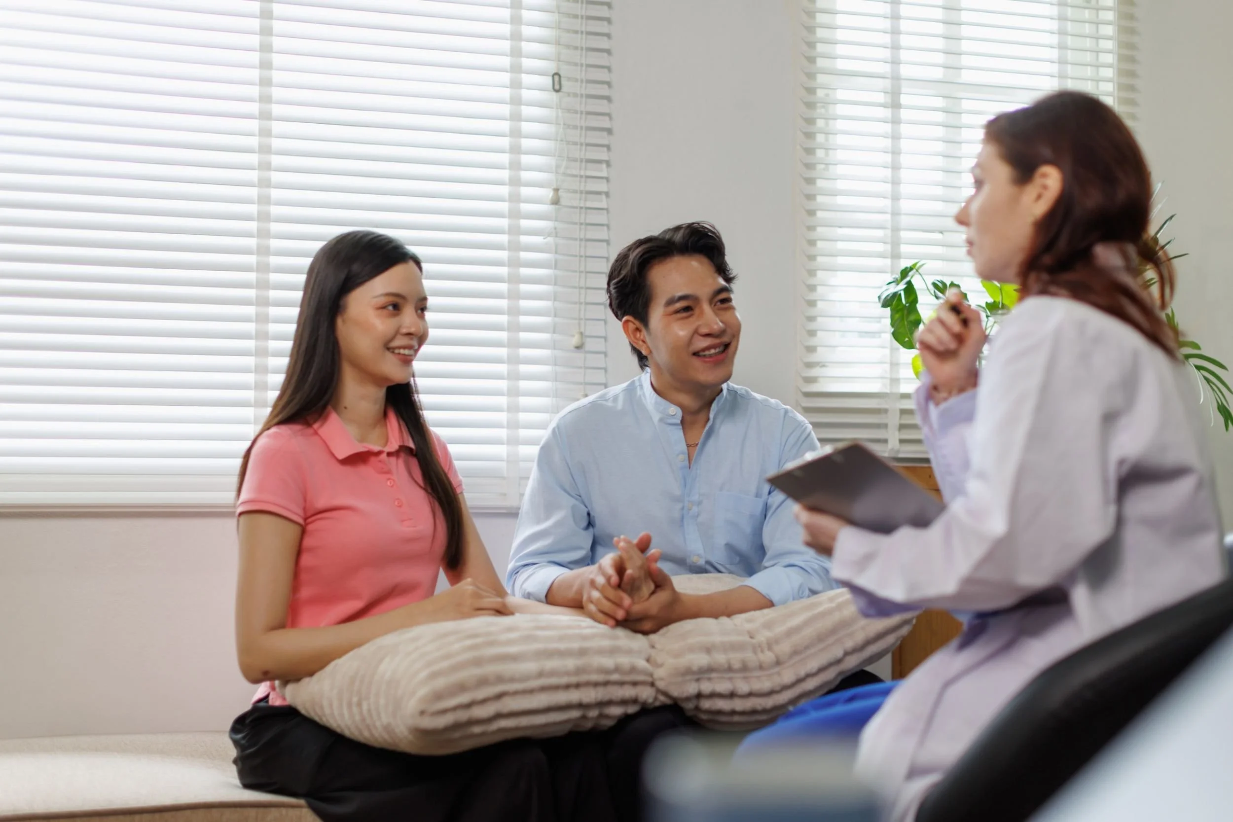 A smiling couple sits together in a clinic, speaking with a doctor in a lab coat holding a clipboard.
