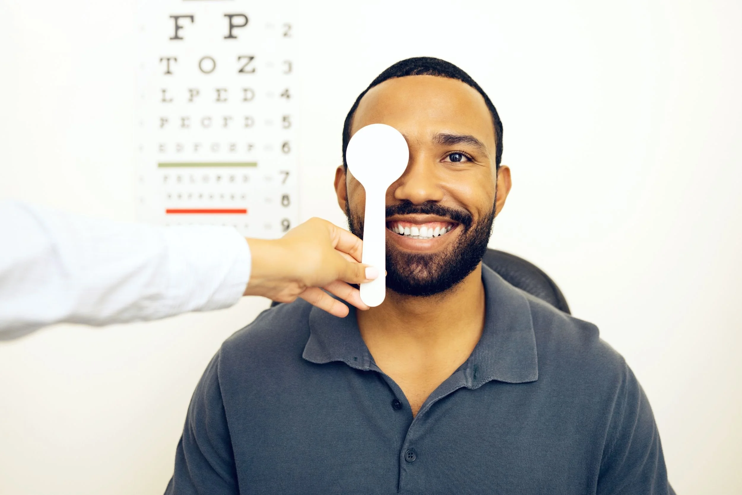 A smiling man sitting in a clinic taking a vision exam while the physician holds a paddle up to one eye.