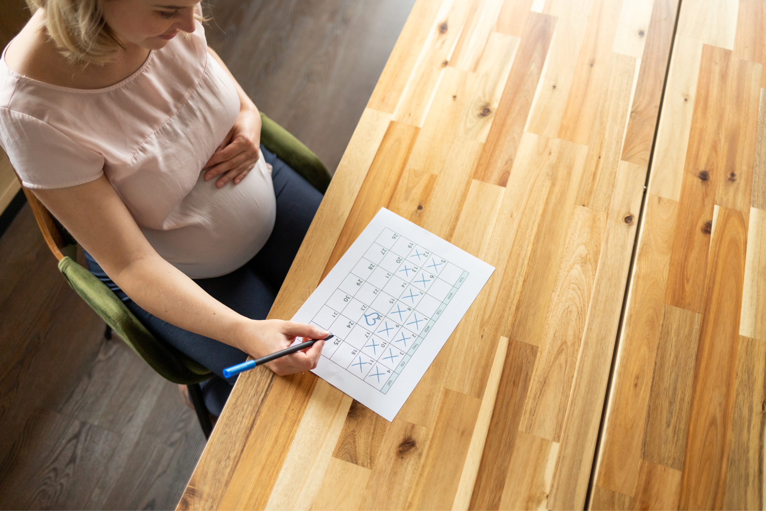 A blonde pregnant woman is sitting at a table with a calendar in front of her, crossing off the days until her due date.