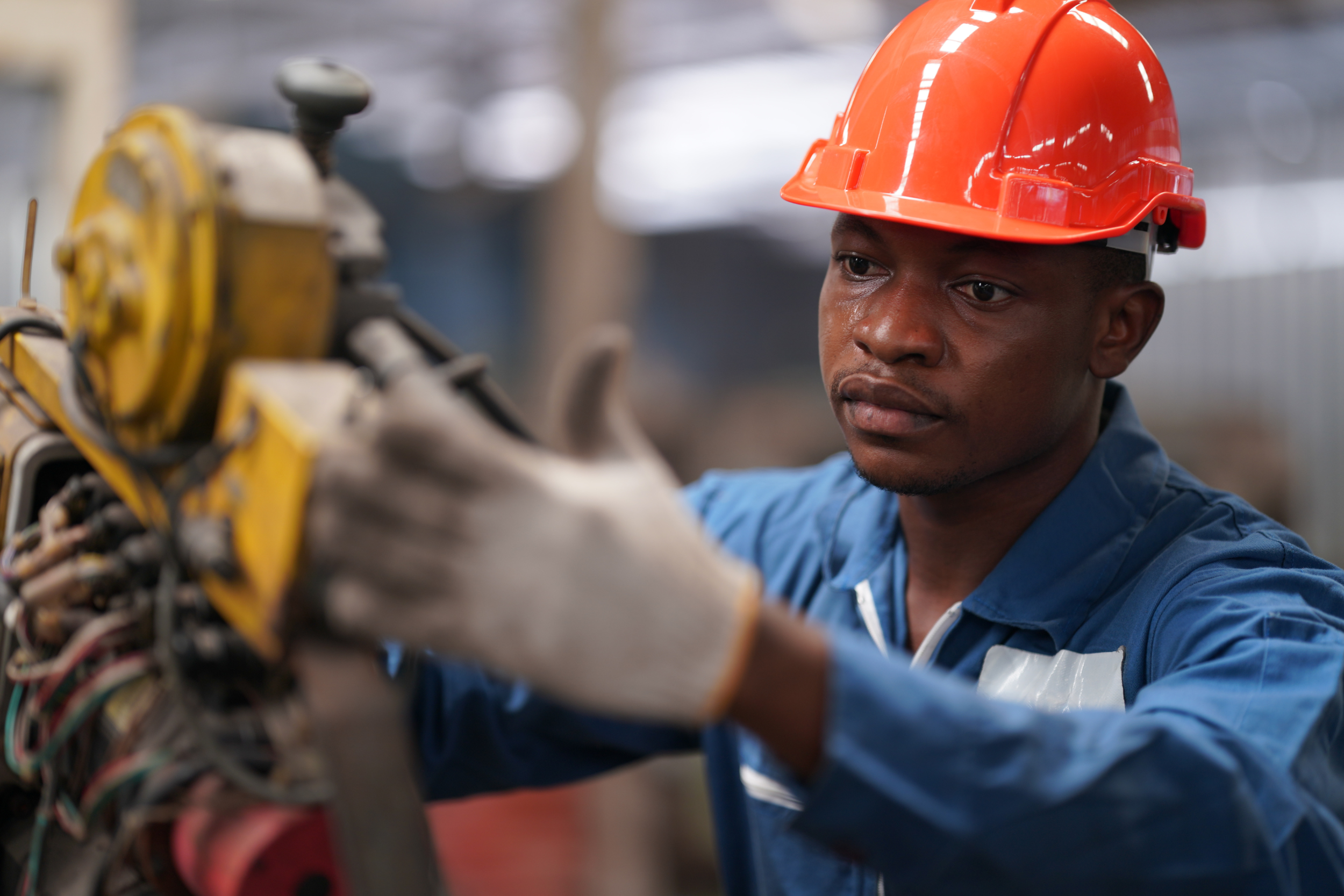 An immigrant skilled trade worker wearing an orange hard hat and blue coveralls, operating machinery.