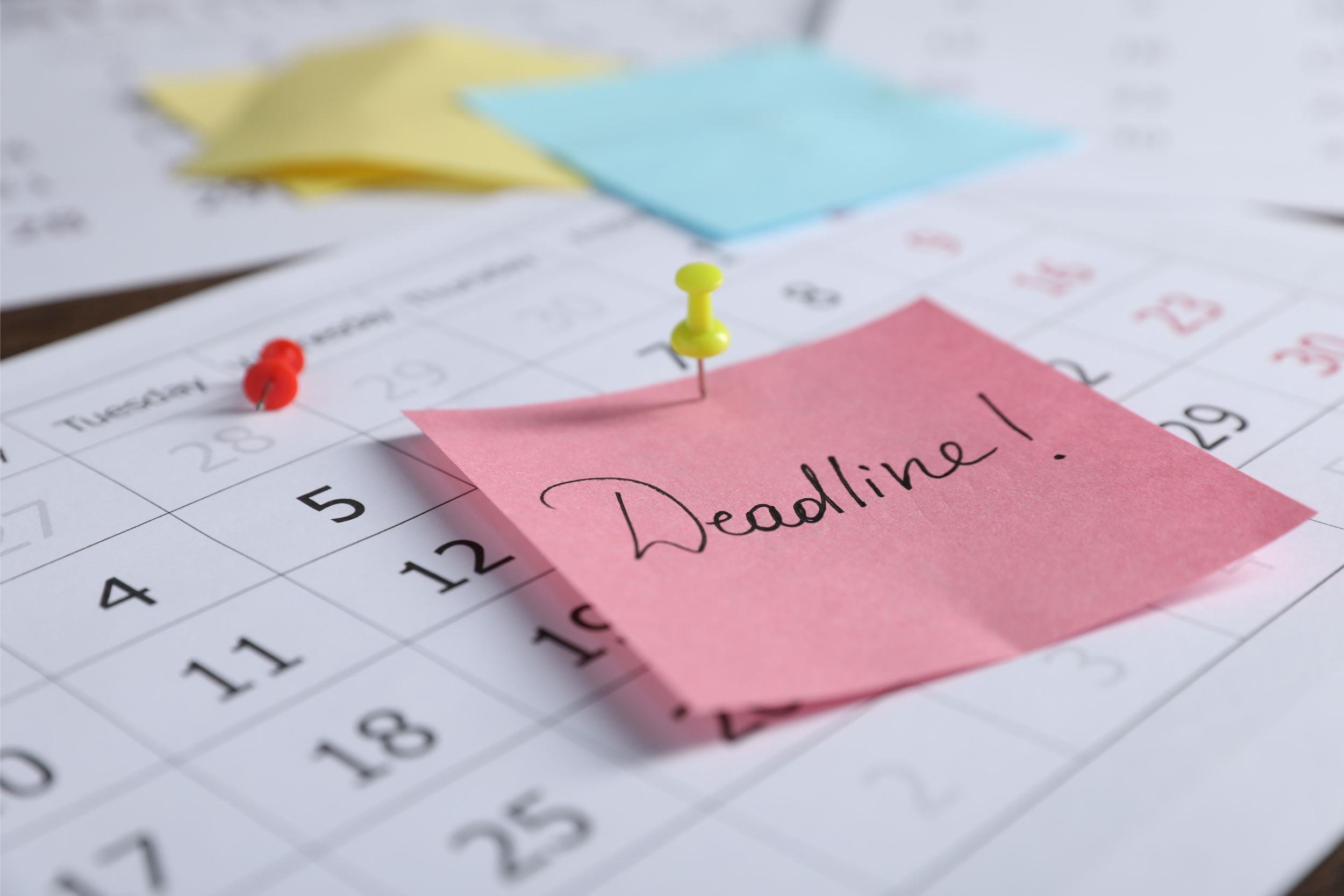 A calendar with a yellow thumbtack pinning a small piece of pink paper with "Deadline!" written on it to a calendar.