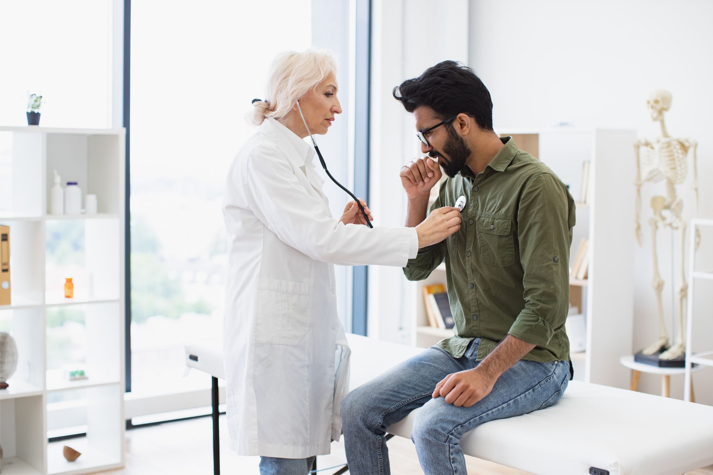 A man sits on a clinic bed and coughs while a doctor in a lab coat listens to his lungs with a stethoscope.