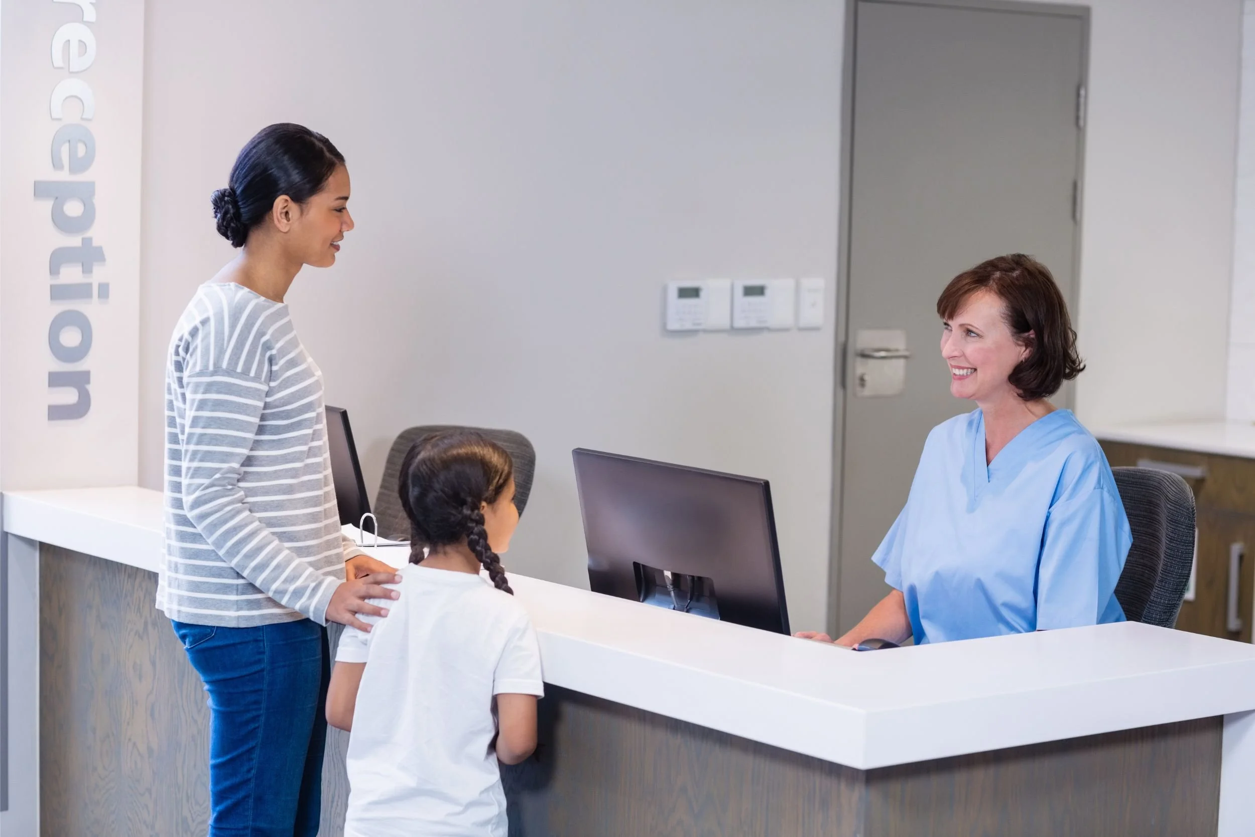 A mother and daughter check in at the reception desk of a clinic, while a smiling nurse checks them in.