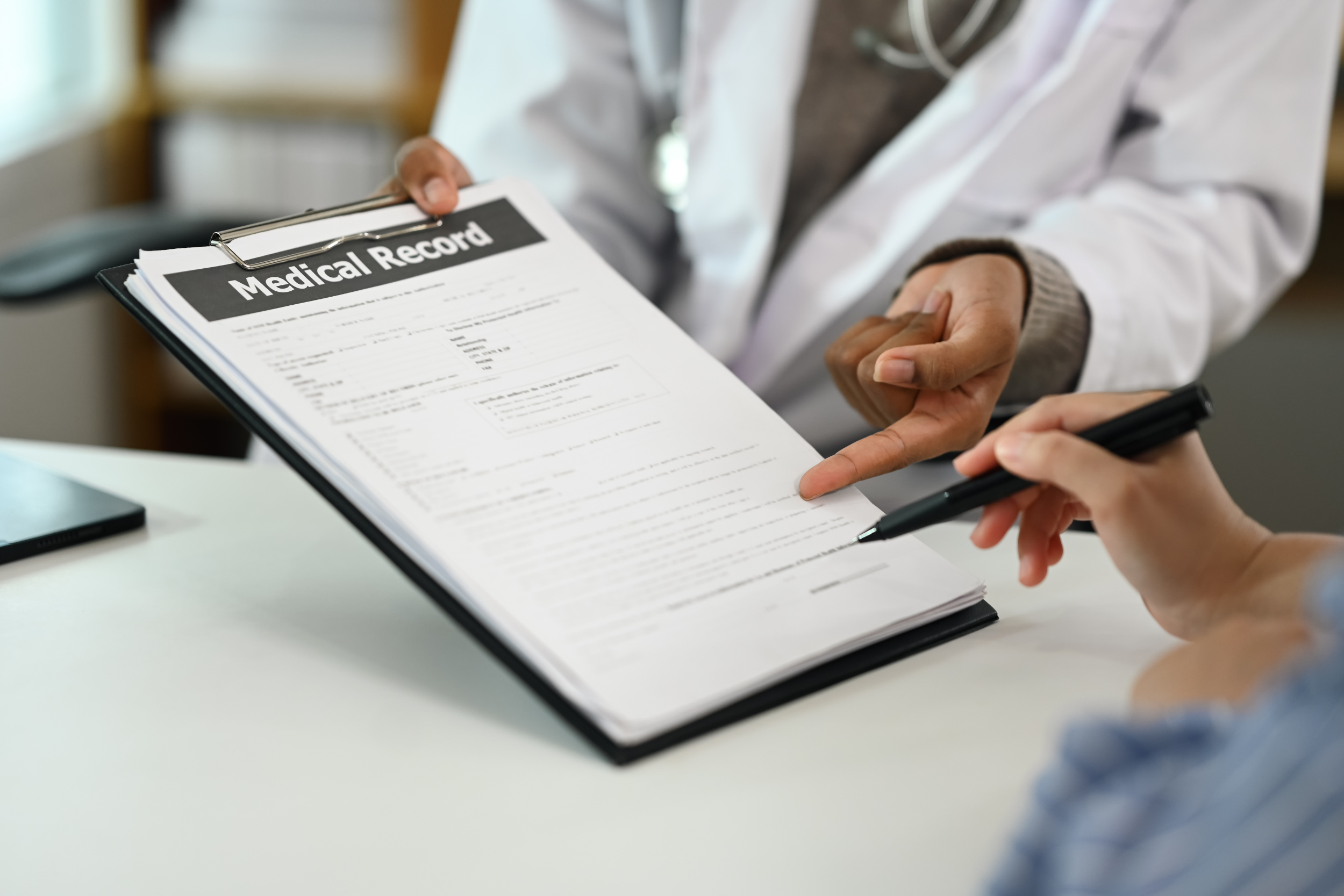 A doctor in a white coat reviews a medical report on a clipboard with a patient who has a pen in hand.