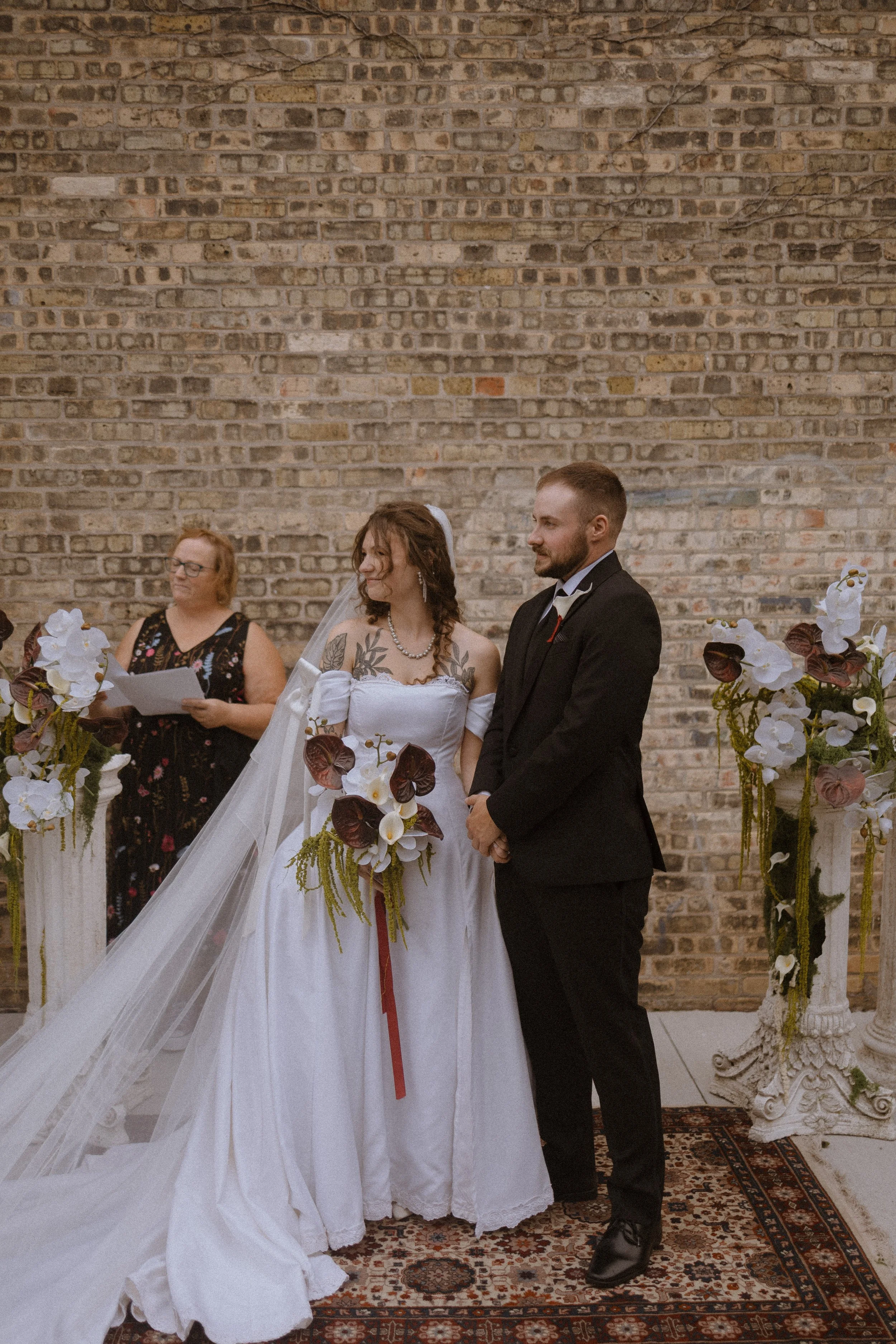 An outdoor wedding ceremony with a bride and groom standing in front of a brick wall, surrounded by floral arrangements, with a woman officiating wearing glasses and holding papers.