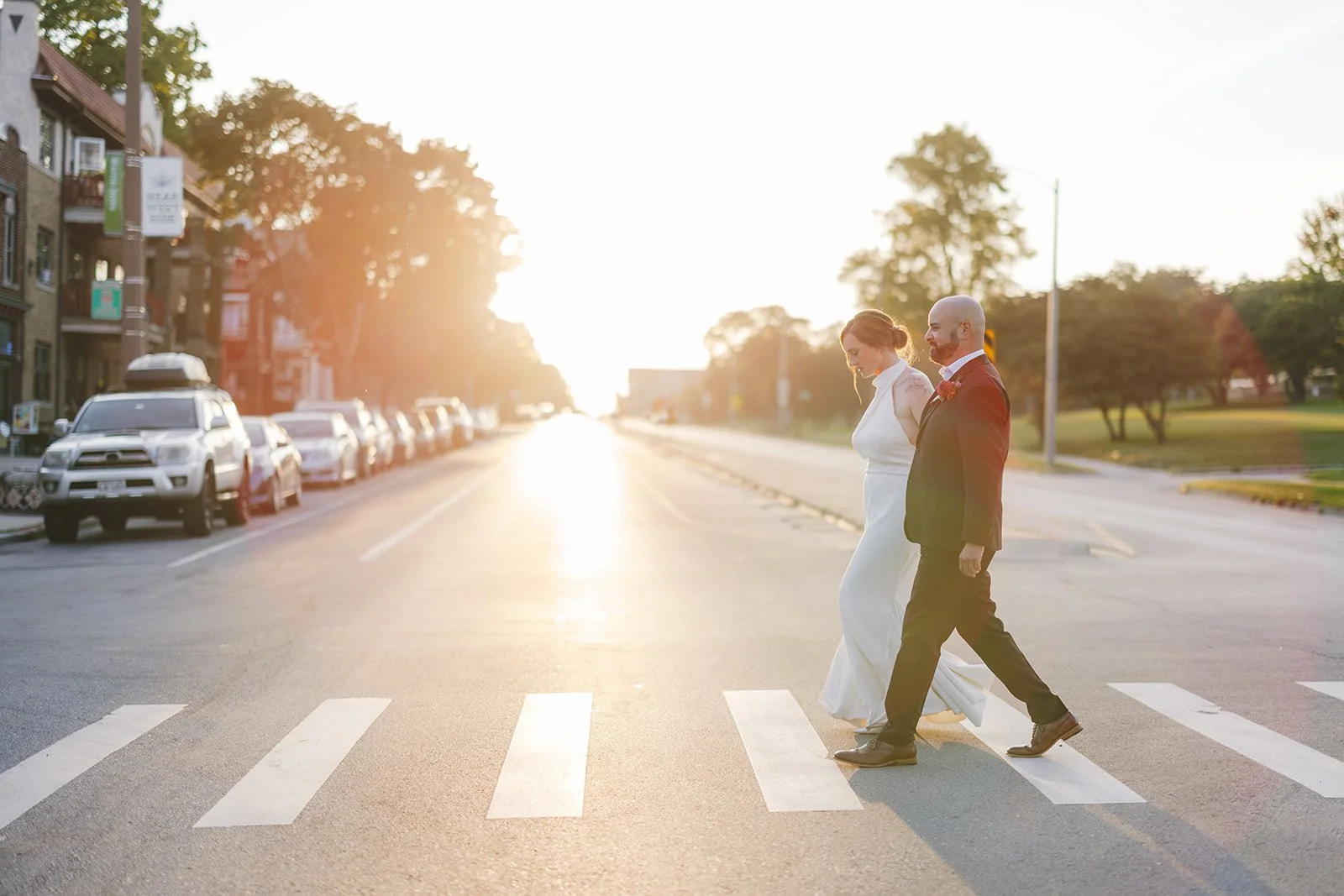 A bride and groom walking through a calm neighborhood, hand in hand across a crosswalk on a city street during sunset, with parked cars and trees in the background.