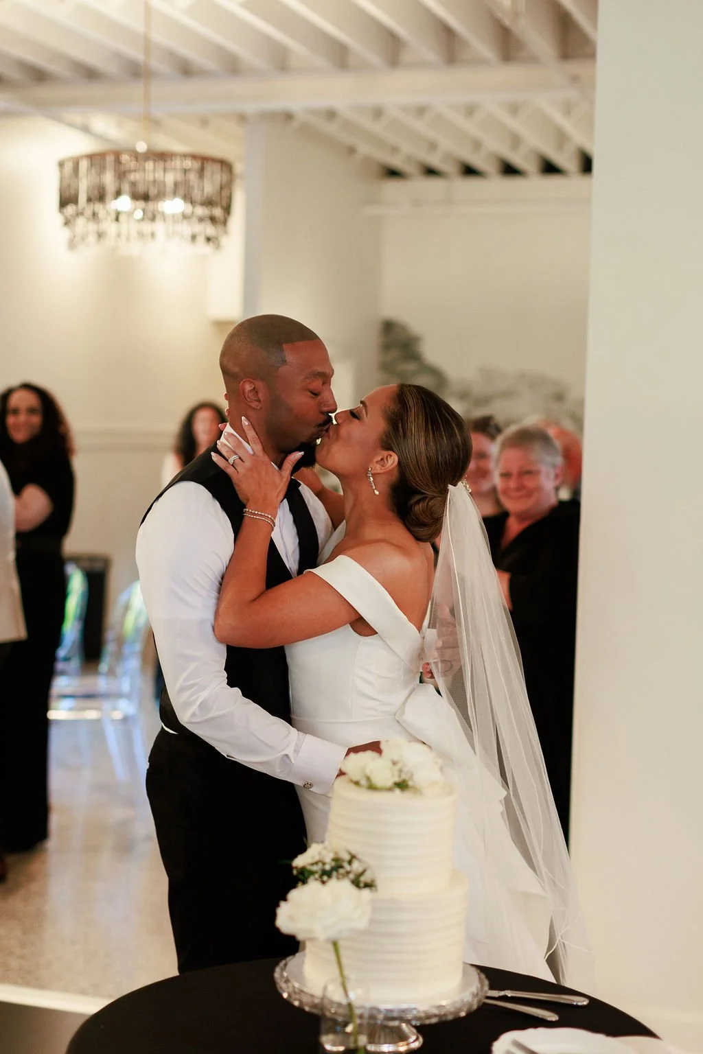 A bride and groom kiss during their wedding reception, with a wedding cake on the table in front of them and guests in the background.