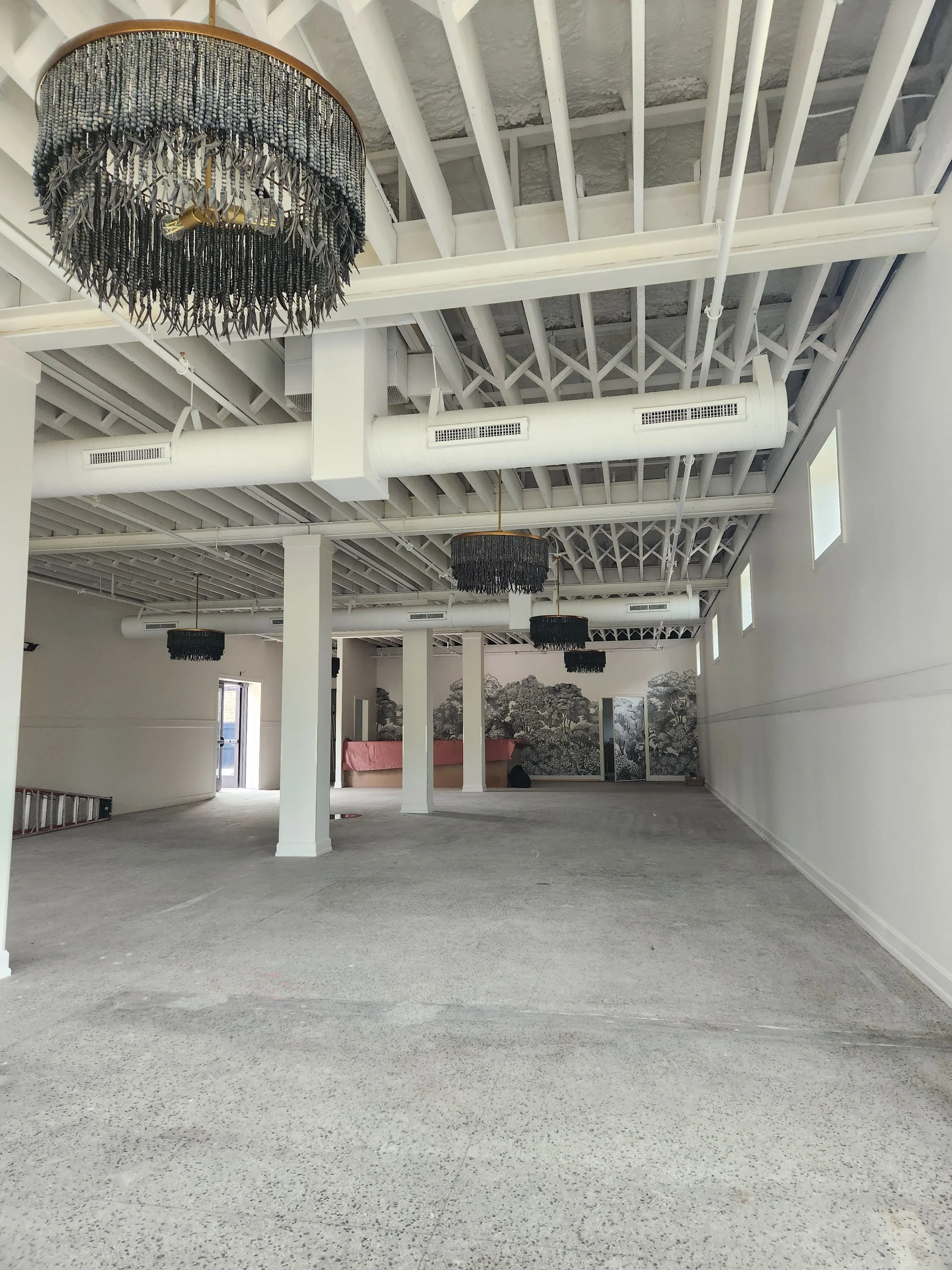 An empty commercial or industrial space with high white walls, exposed ceiling beams, and hanging black chandeliers with beaded fringe. Large windows allow natural light to fill the room, and there is a mural of trees on the far wall.