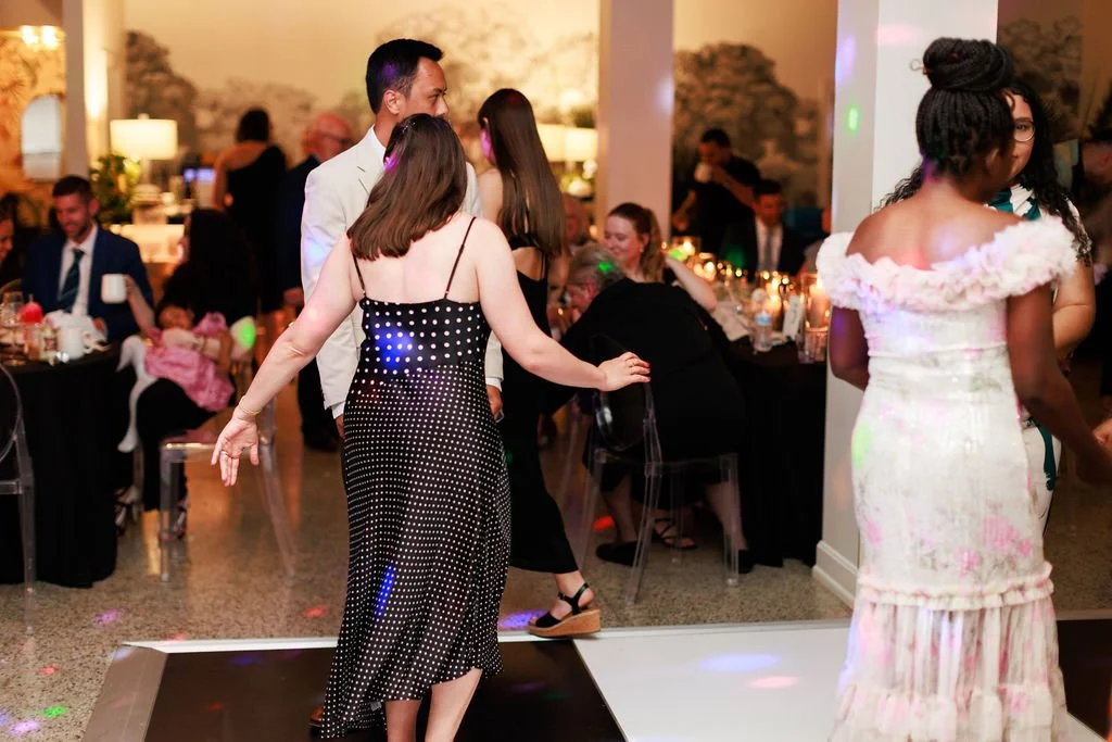 People dancing and socializing at a party or celebration, with some seated at tables in the background.