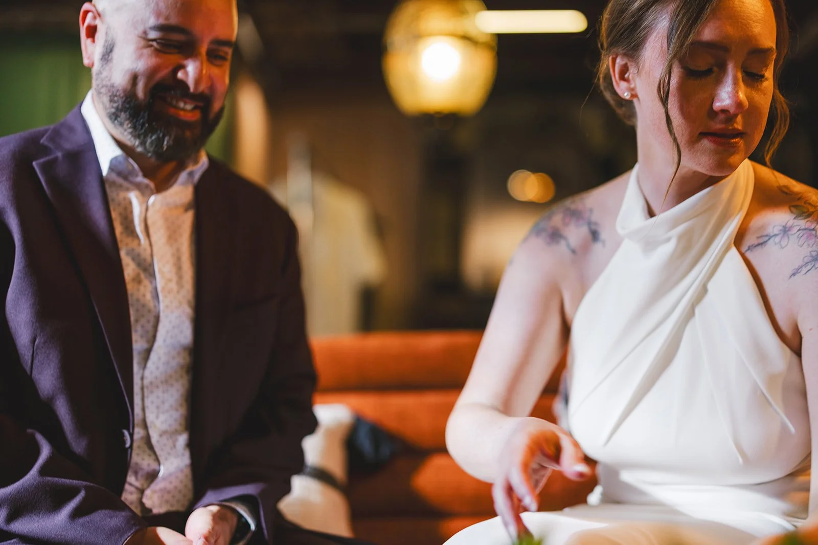 A man with a beard wearing a suit and a woman with tattoos wearing a white dress sitting together in a warmly lit room.