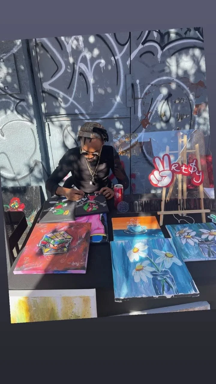 An artist sitting at a table displaying colorful paintings including a Rubik's cube, a mug, and flowers. The background has graffiti art, and a sign with a peace hand and the words 'Metchy' is visible.