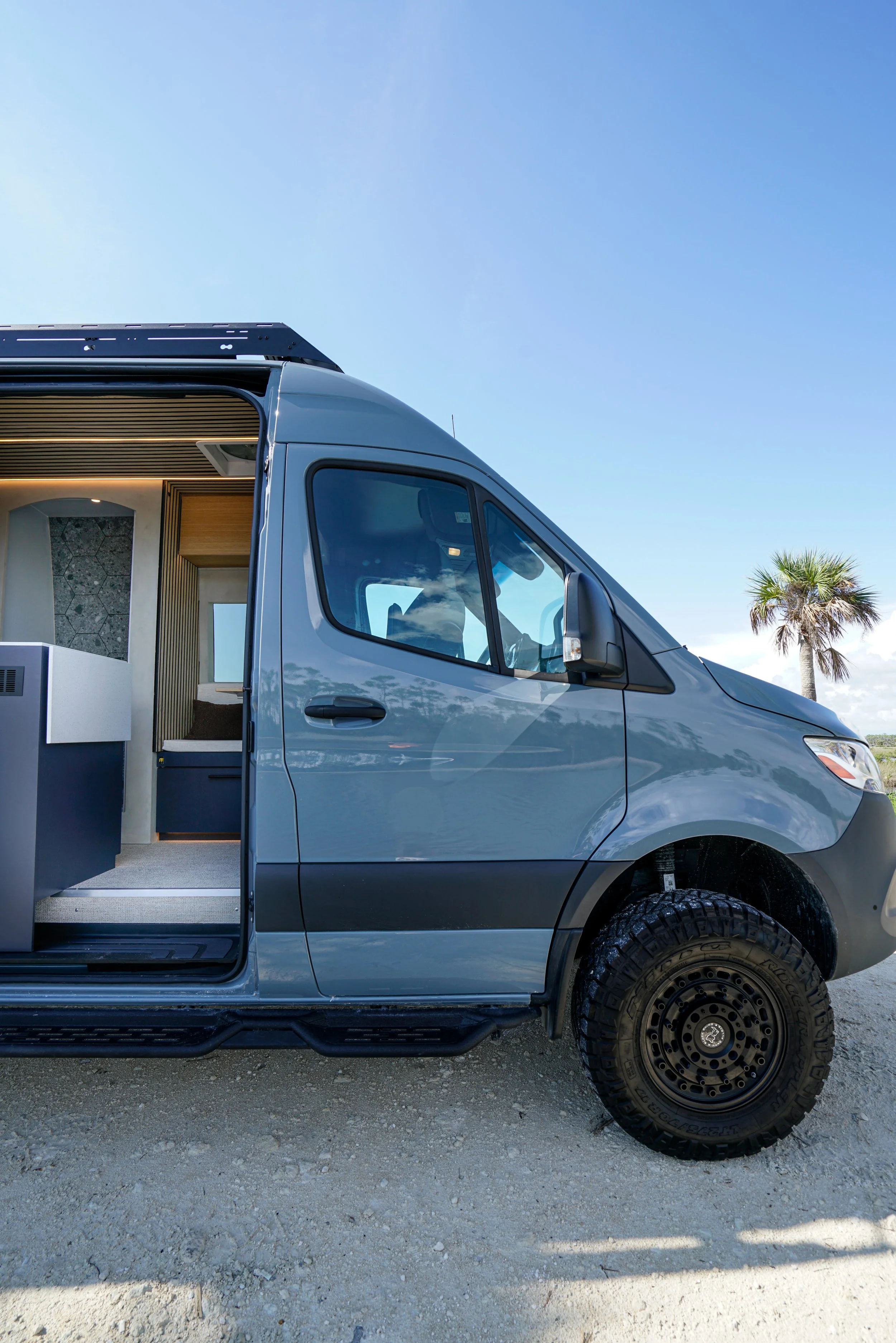 Side view of a gray camper van with a partially open door, showing a modern interior with wooden accents and hexagon-patterned wall, parked outdoors on a gravel surface under a clear blue sky with a palm tree in the background.