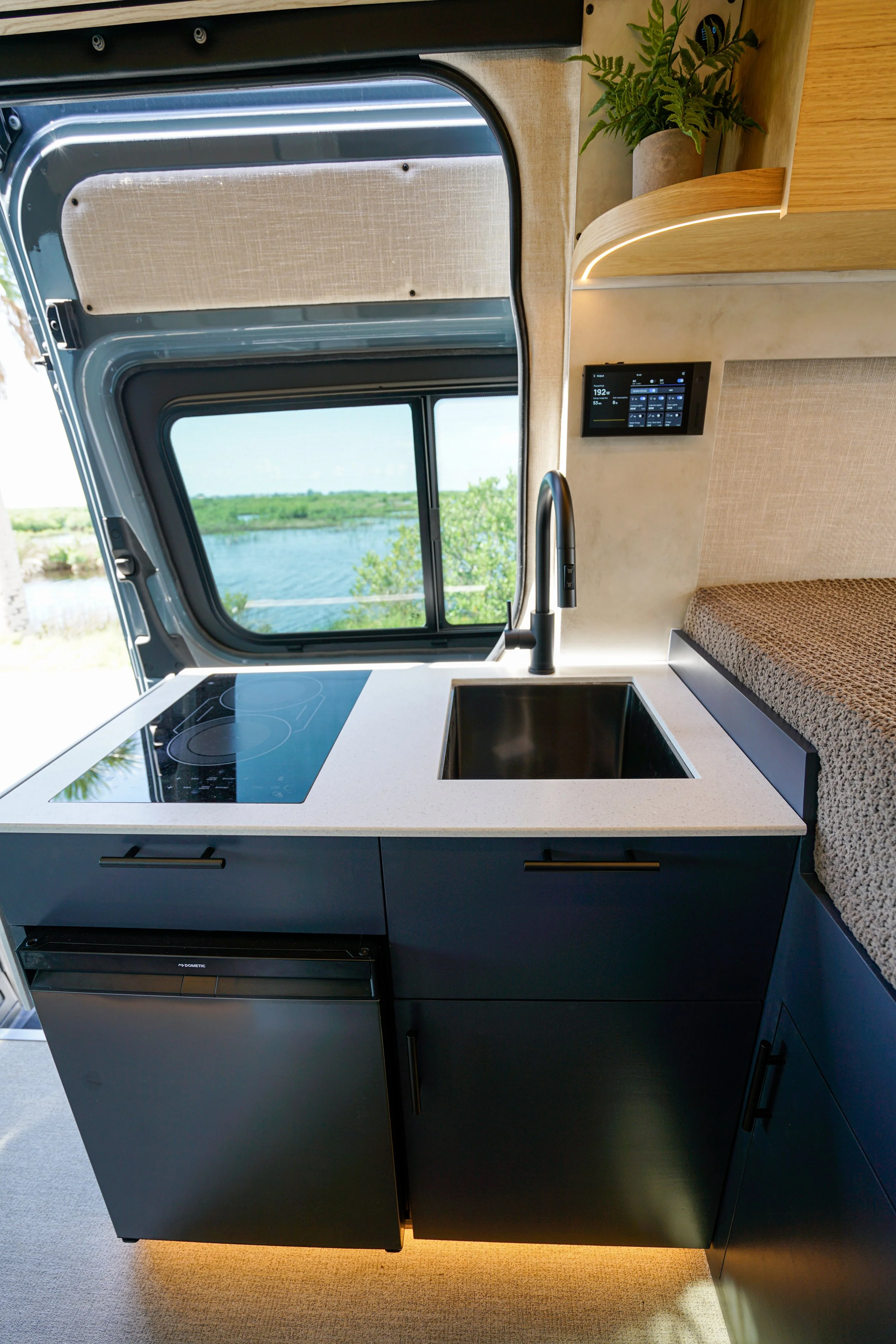 Kitchen area inside a camper van with a black induction cooktop, a sink, and a view of water and greenery through a large window.